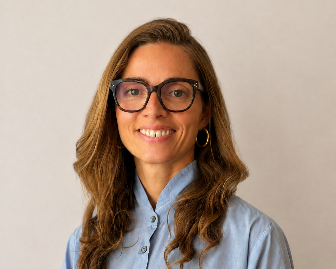 Porträt einer lachenden Frau mit langem braunem Haar, trägt schwarze Brille, goldene Ohrringe und eine blaue Bluse vor hellem Hintergrund.