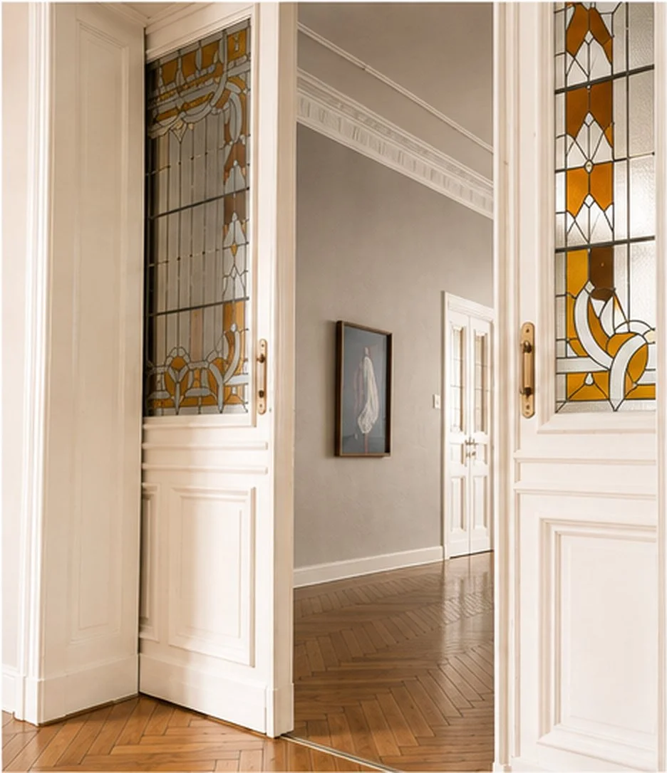 Blick in einen Flur mit Holzboden und knarrende Dielen, durch eine halbgeöffnete Tür aus Glas mit dekorativen Mustern, davor eine weiße Tür mit goldener Türklinke, Wandfarbe in Grau, ein eingerahmtes Bild an der Wand.