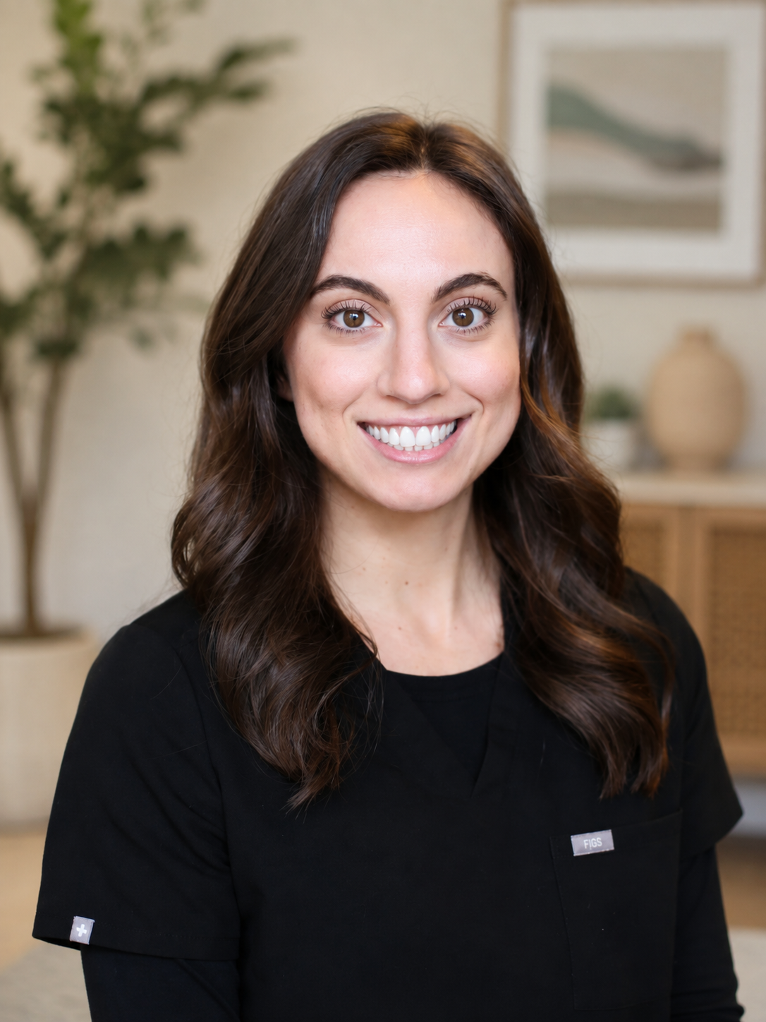 A smiling woman with long brown hair wearing a black shirt standing indoors with a plant, framed artwork, and decorative objects in the background.