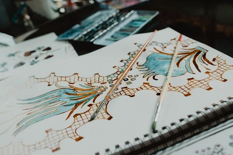 Artistic design on paper with watercolor painting of a blue vase with flowers, surrounded by intricate orange patterns, with paintbrushes resting on it.