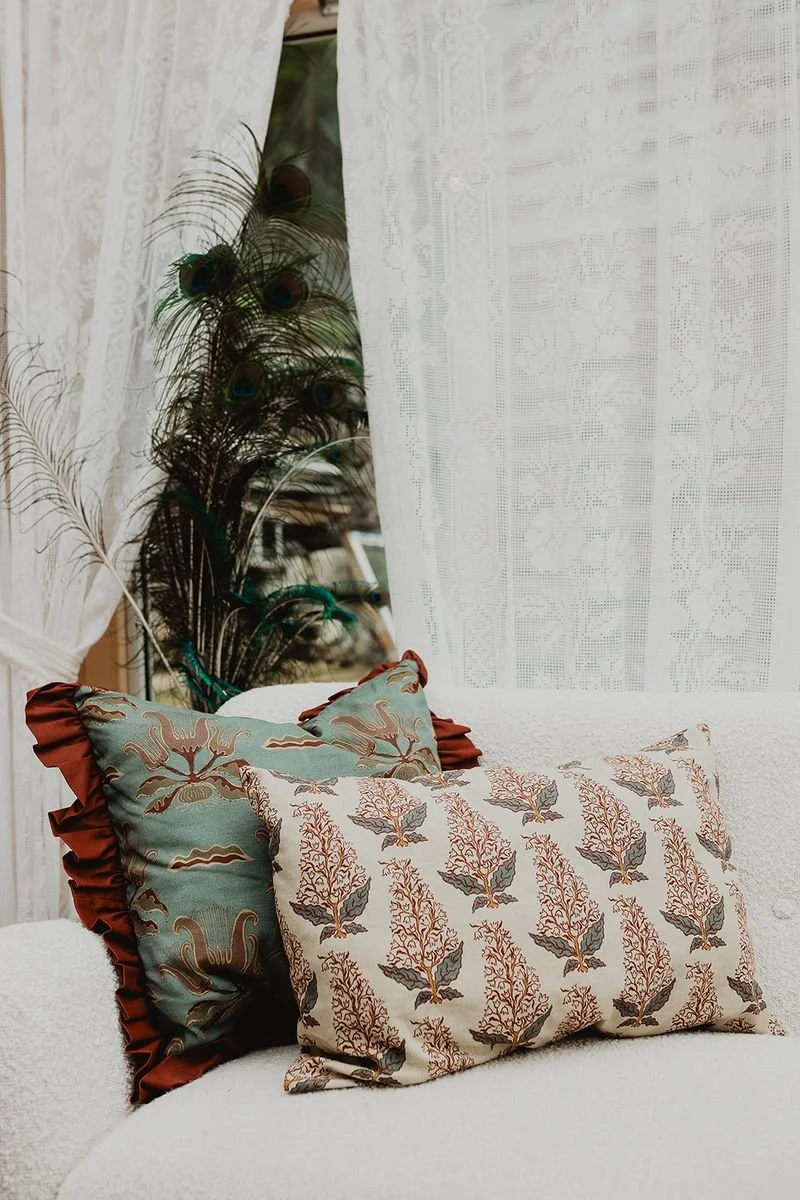 Decorative pillows on a white couch, with peacock feathers and lace curtains in the background.
