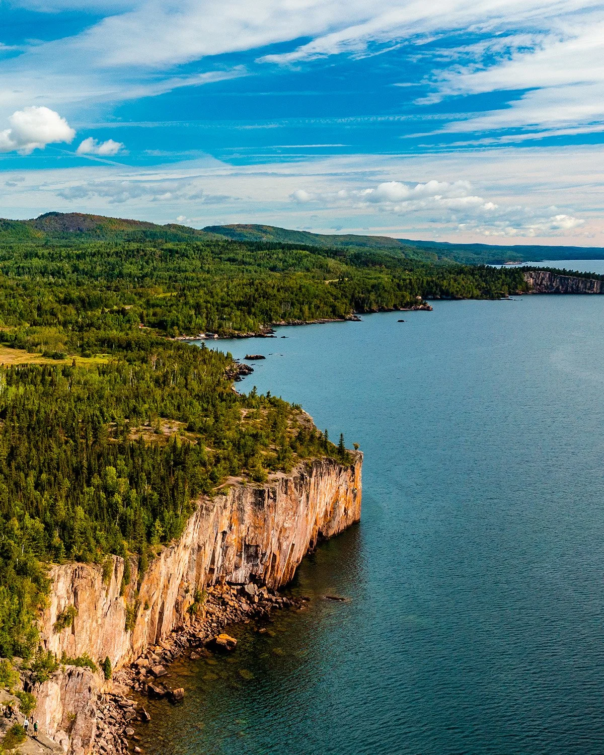 Scenic view of a large body of water with steep cliffs covered in dense green trees along the shoreline. In the background, rolling hills and a partly cloudy sky with streaks of white clouds.