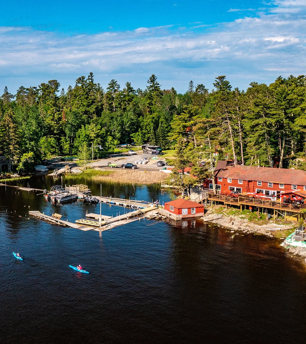 Aerial view of a lakeside area with boats docked at a marina, a red wooden building with an outdoor deck, surrounded by tall green trees, with a parking lot in the background under a blue sky with some clouds.