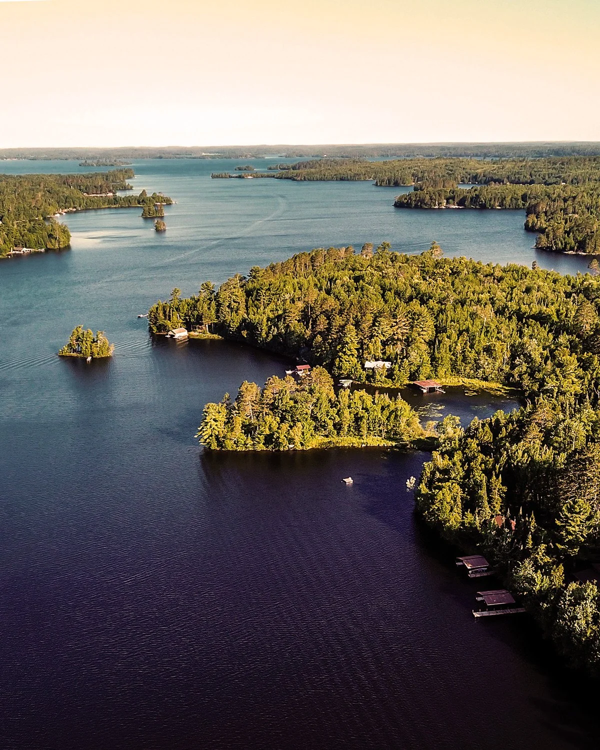 Aerial view of a large lake with numerous small islands covered in green trees, some with houses and docks along the shore, extending into the distance under a clear sky.