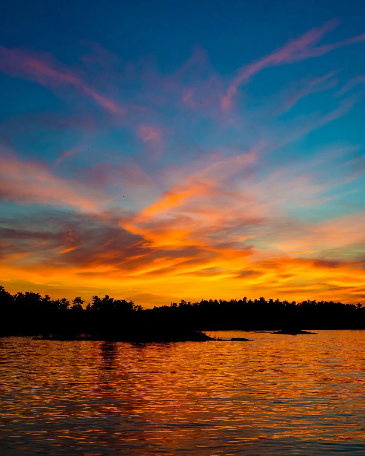 A vibrant sunset over a body of water with colorful orange, yellow, pink, and blue clouds and a silhouette of trees in the distance.