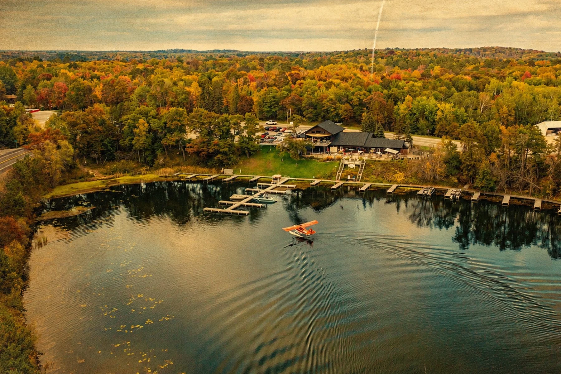 Aerial view of a lake with a seaplane taking off, surrounded by autumn-colored trees and a building near the shore with docks and boats.