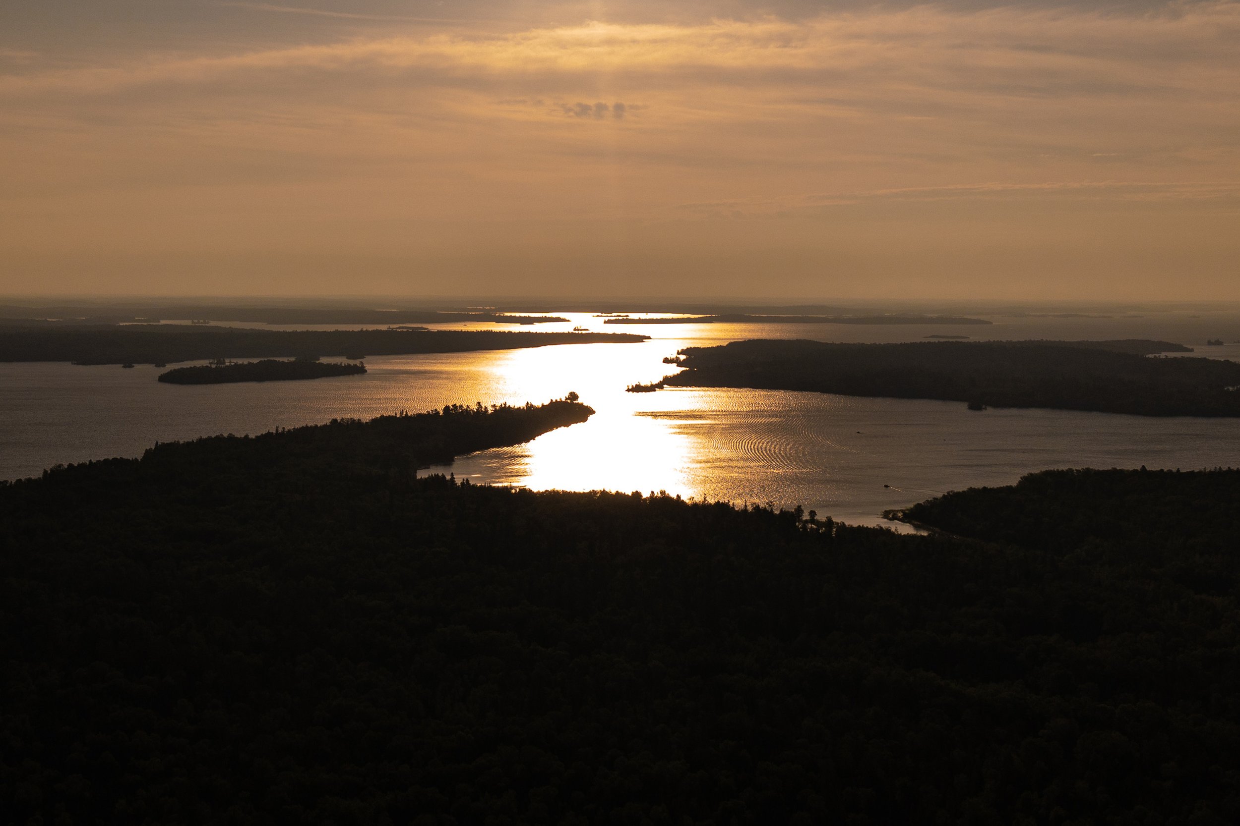 A scenic sunset over a body of water with islands and landmasses, casting a reflective glow on the water surface and a sky with scattered clouds.