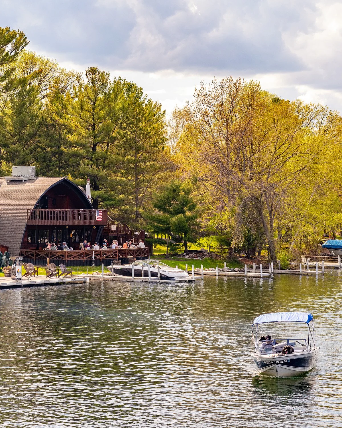 A lakeside scene with a boat on the water, a dock with piers, and a house surrounded by trees with green and yellow leaves. People are sitting outside on a deck of the house.