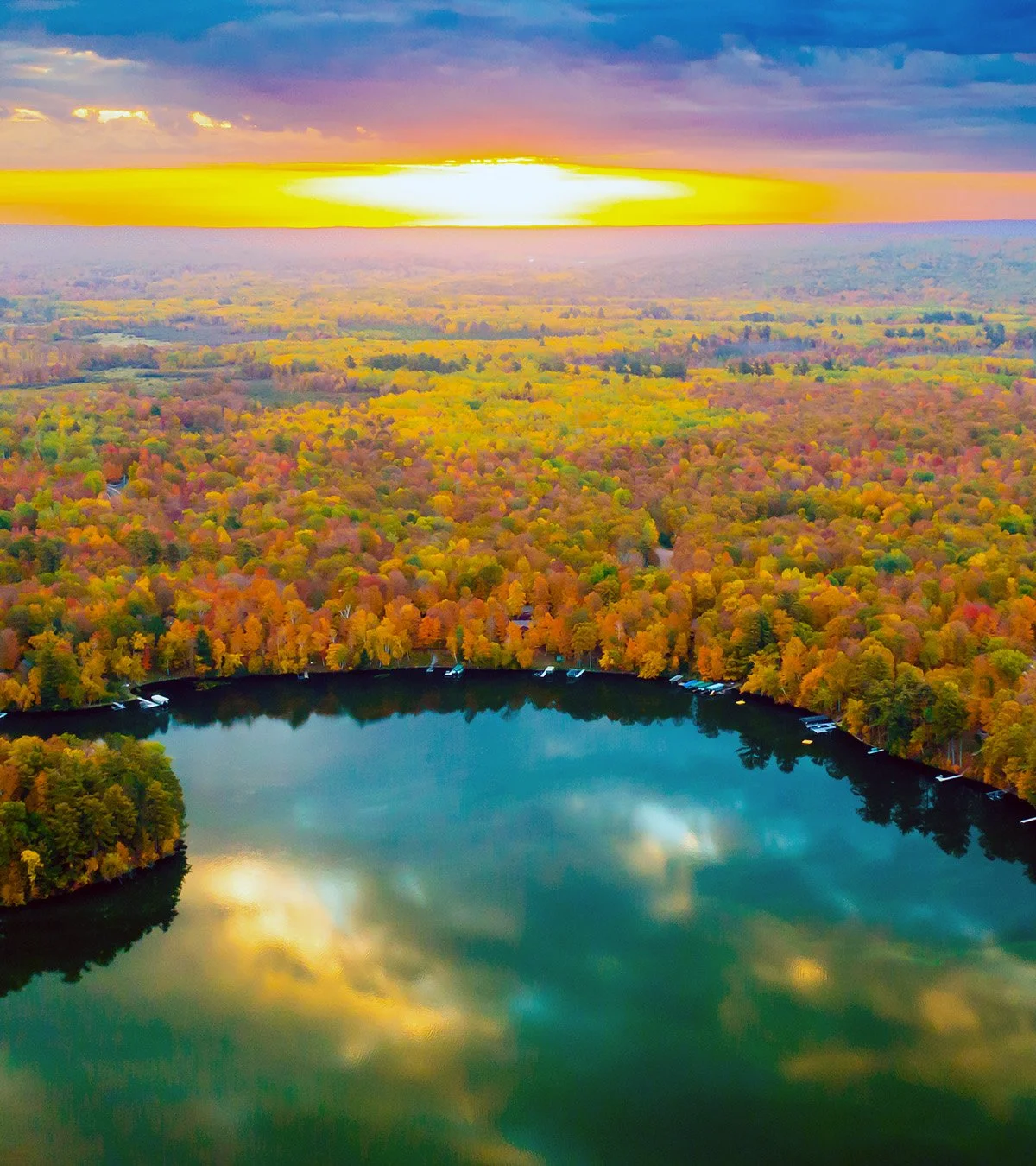 An aerial view of a lake surrounded by a forest with trees displaying fall colors, under a sky with clouds and a setting sun casting a golden glow.