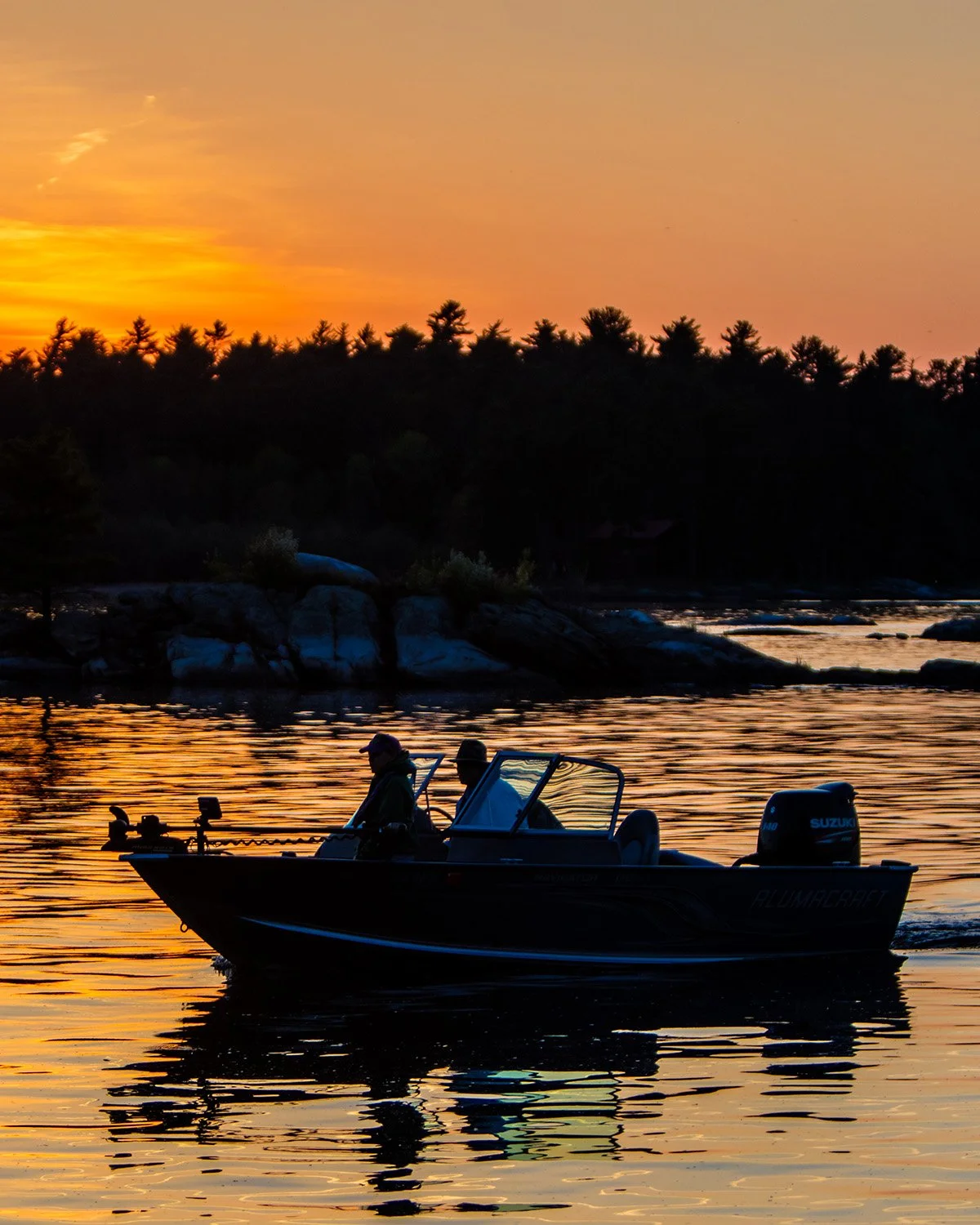 A boat with two people on a lake during sunset, with the sky in shades of orange and pink, and trees along the shoreline in the background.