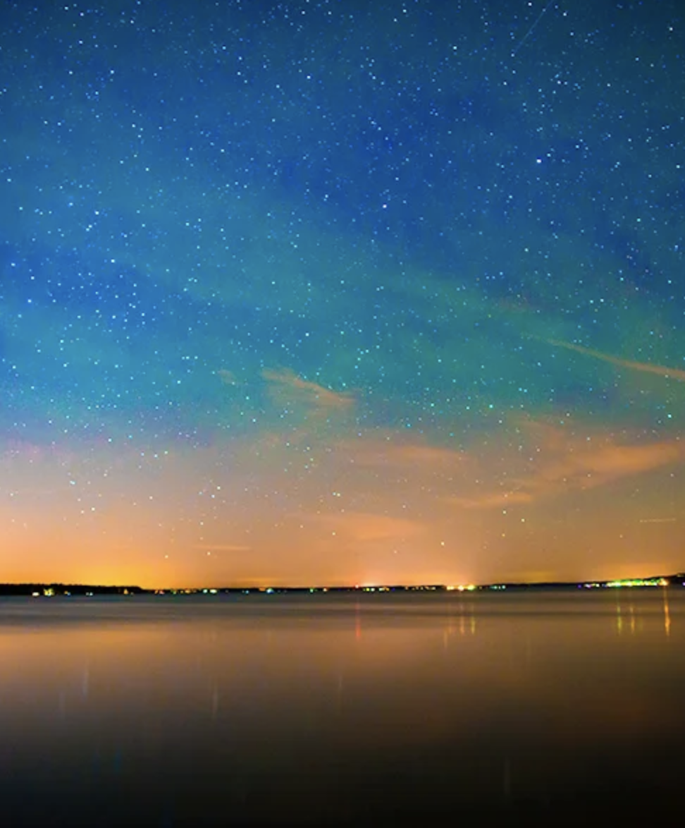Night sky filled with stars over a calm body of water, with a faint glow on the horizon.