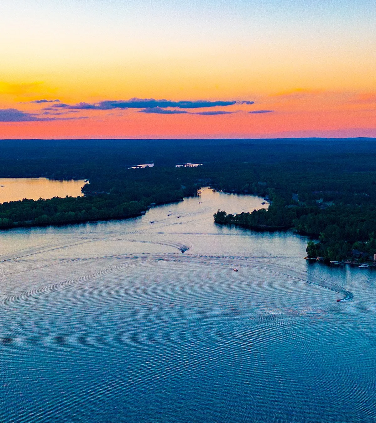 Aerial view of a river during sunset with colorful sky, surrounded by dense green forest and a few boats creating ripples in the water.