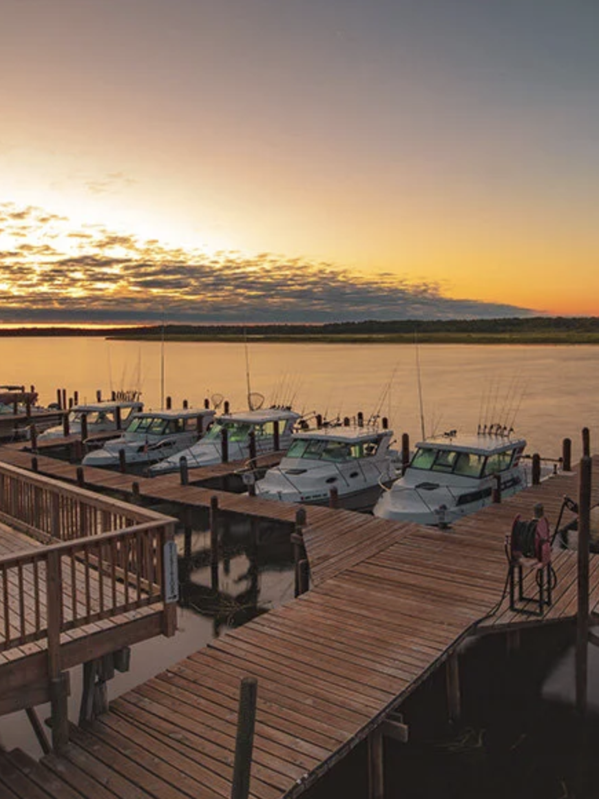 A marina with several white boats docked at wooden piers during sunset, with a calm body of water and a sky filled with clouds and warm colors.
