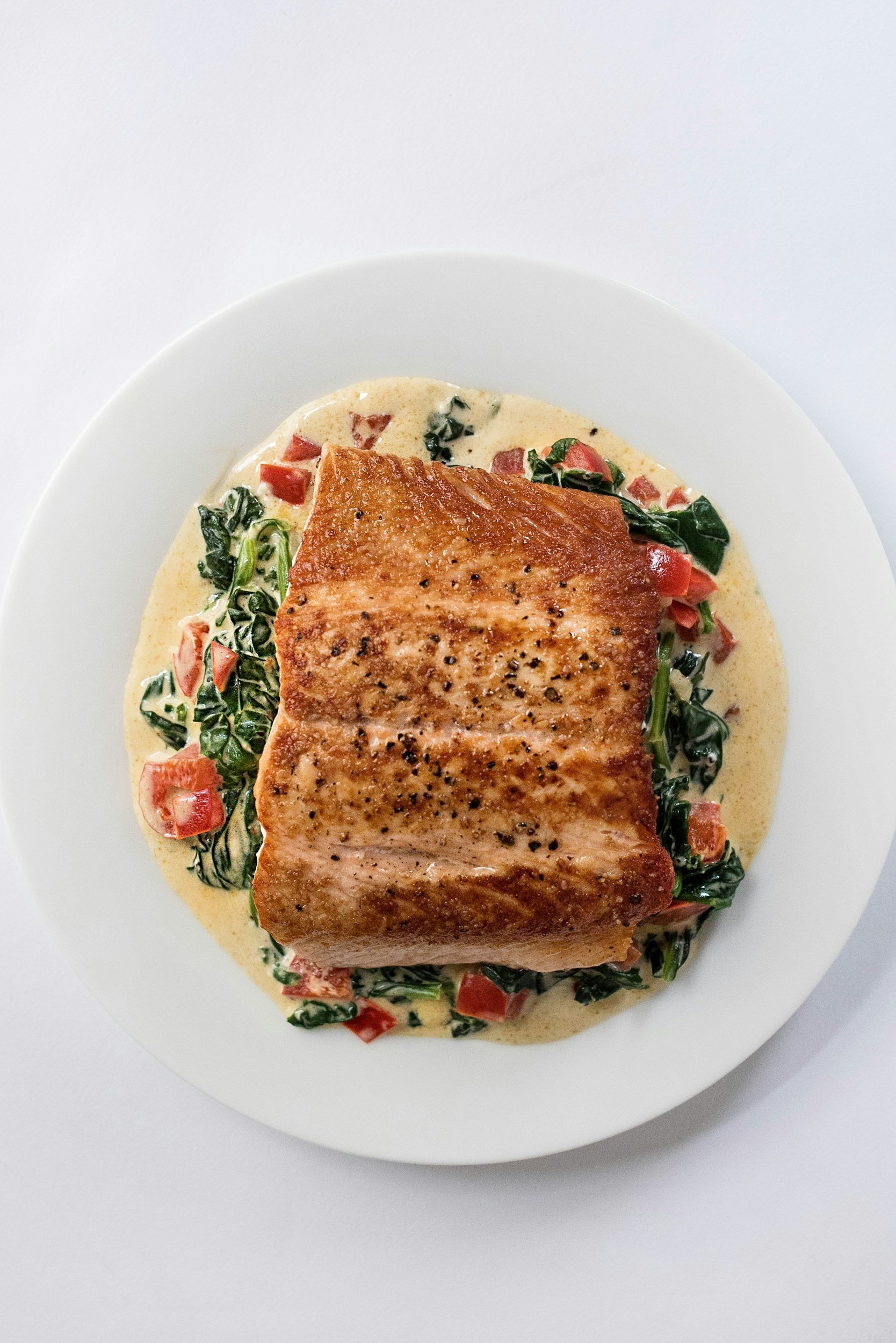 A plate of cooked salmon topped with black pepper, served over a bed of creamy spinach and tomato mixture on a white dish.