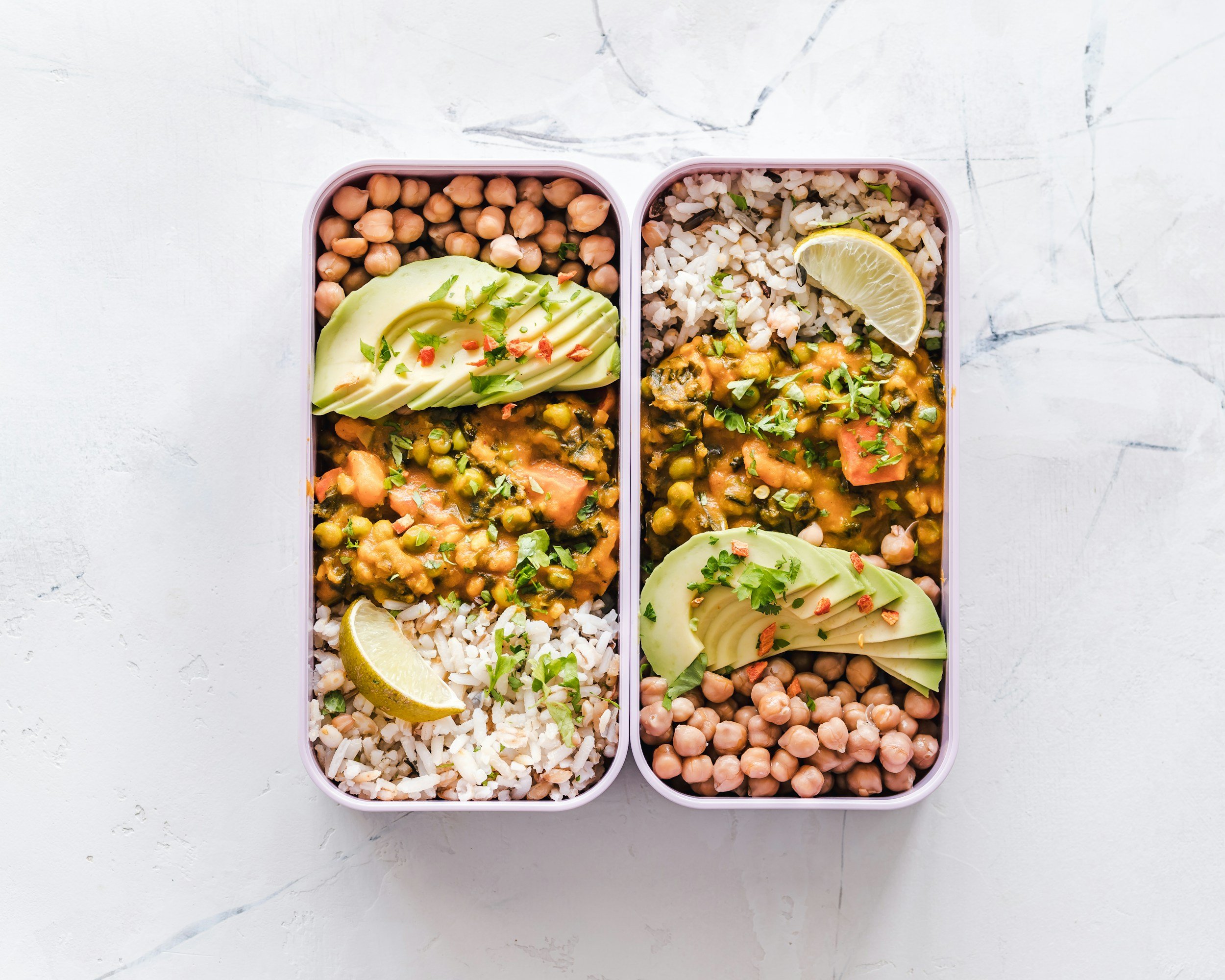 Two meal prep containers filled with chickpeas, brown rice, avocado slices, a lemon wedge, and a vegetable curry garnished with chopped herbs.