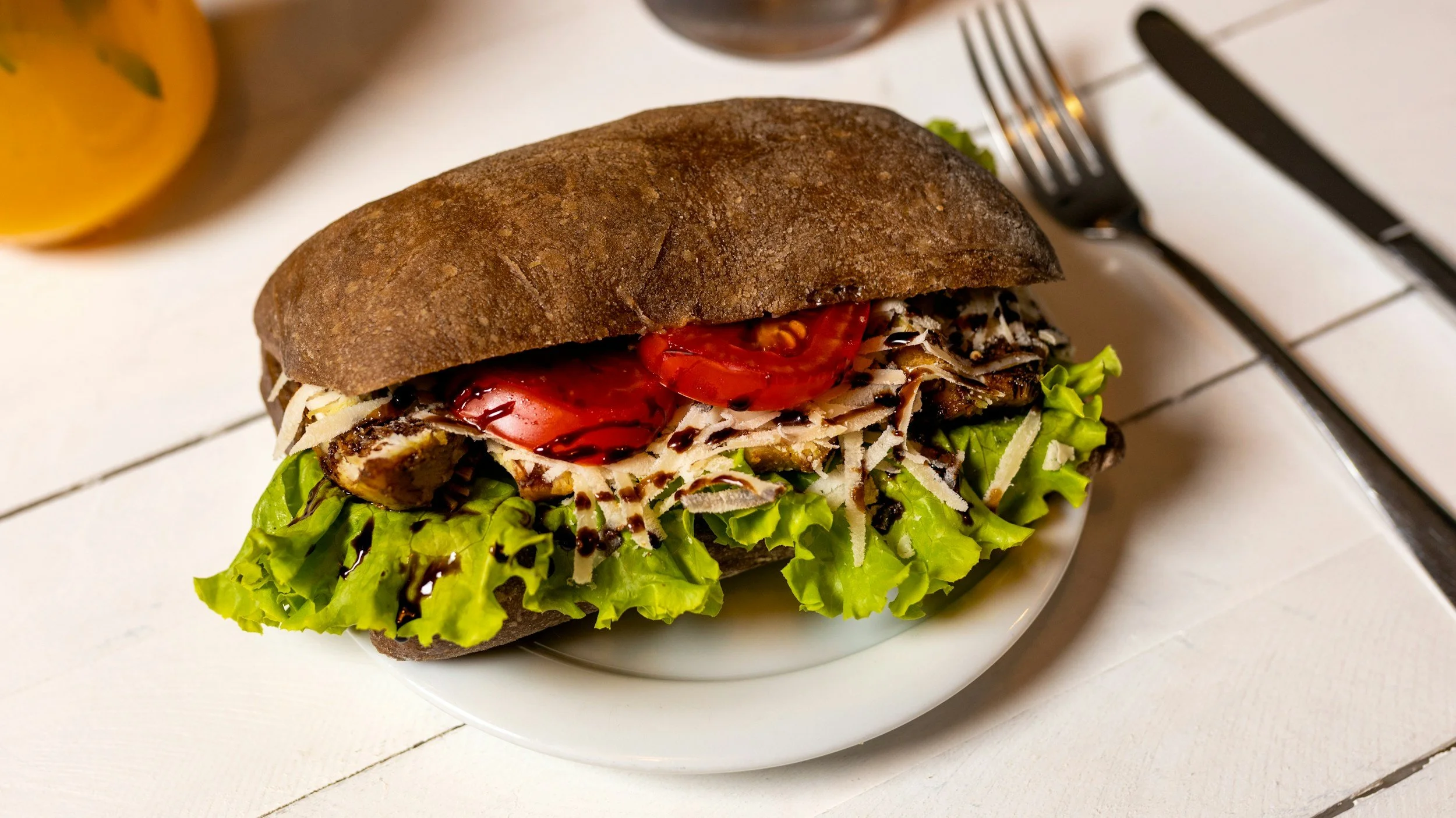 Sandwich on a white plate with lettuce, sliced tomato, shredded cheese, and dark brown sandwich bread on a white table, with a fork and knife to the right and a yellow beverage in the background.