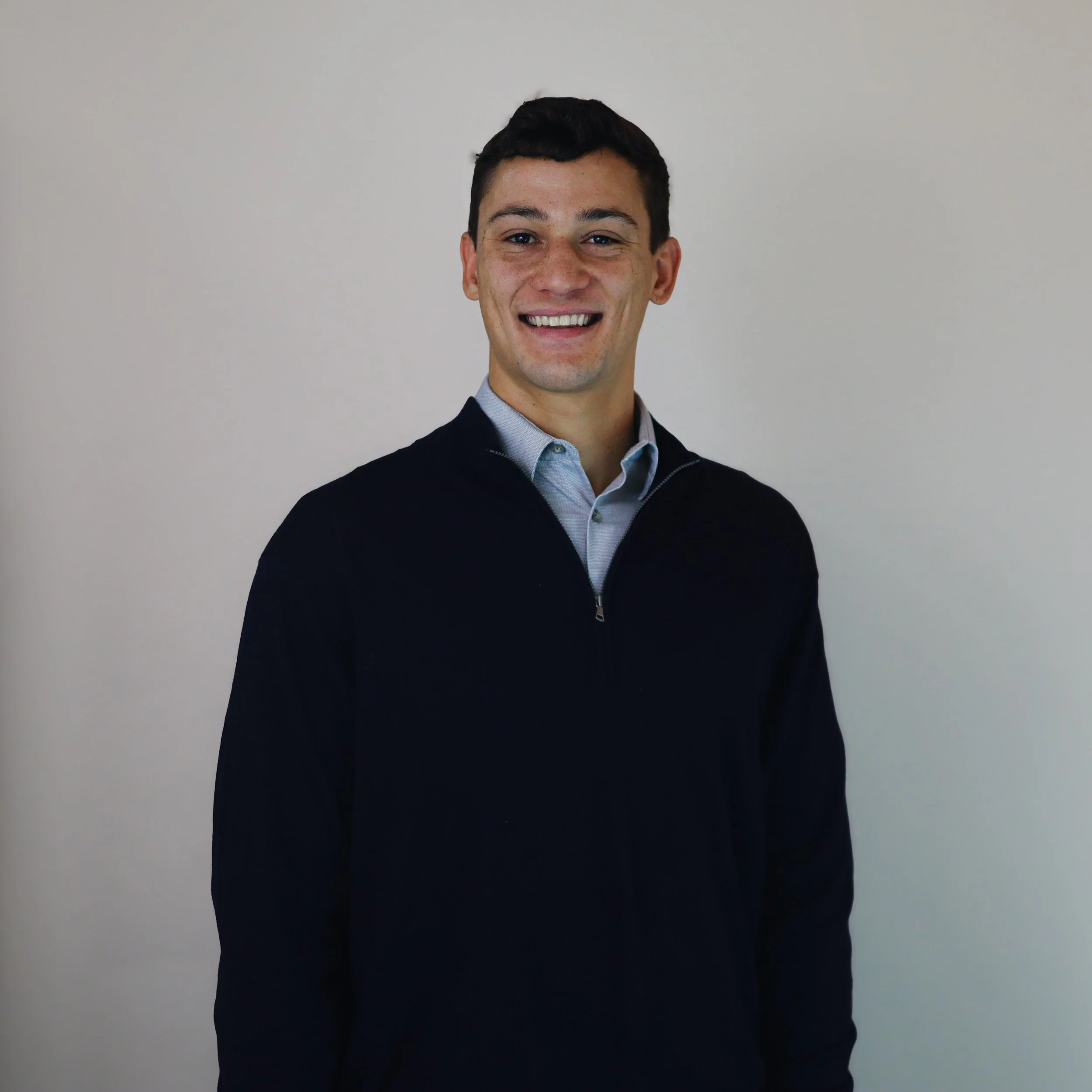 Portrait of a young man with short dark hair, smiling, wearing a collared shirt under a dark zip-up sweater, standing against a plain light-colored wall.