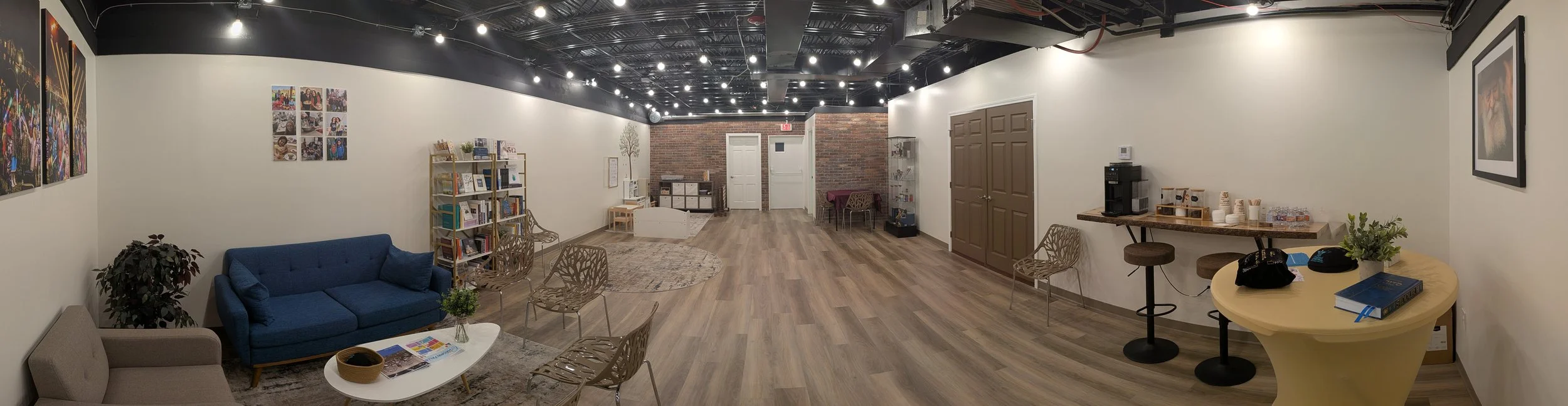 Interior of a community or lounge space with white walls, wood flooring, and an exposed brick section. Furniture includes a beige sofa, a blue sofa, several chairs, and a coffee table with magazines. There are bookshelves, artwork on the walls, a coffee station with cups and a coffee maker, and a small table with a plant and books. Overhead lights illuminate the room.