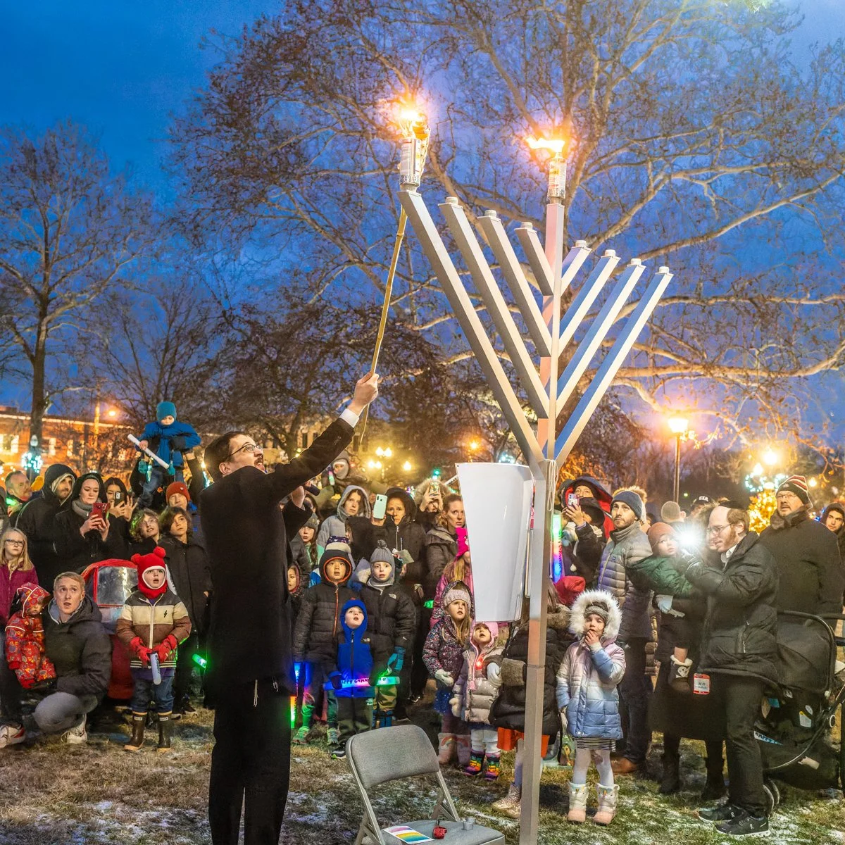 People gathered outdoors in the evening for a Hanukkah celebration, lighting a large menorah as children and adults watch.