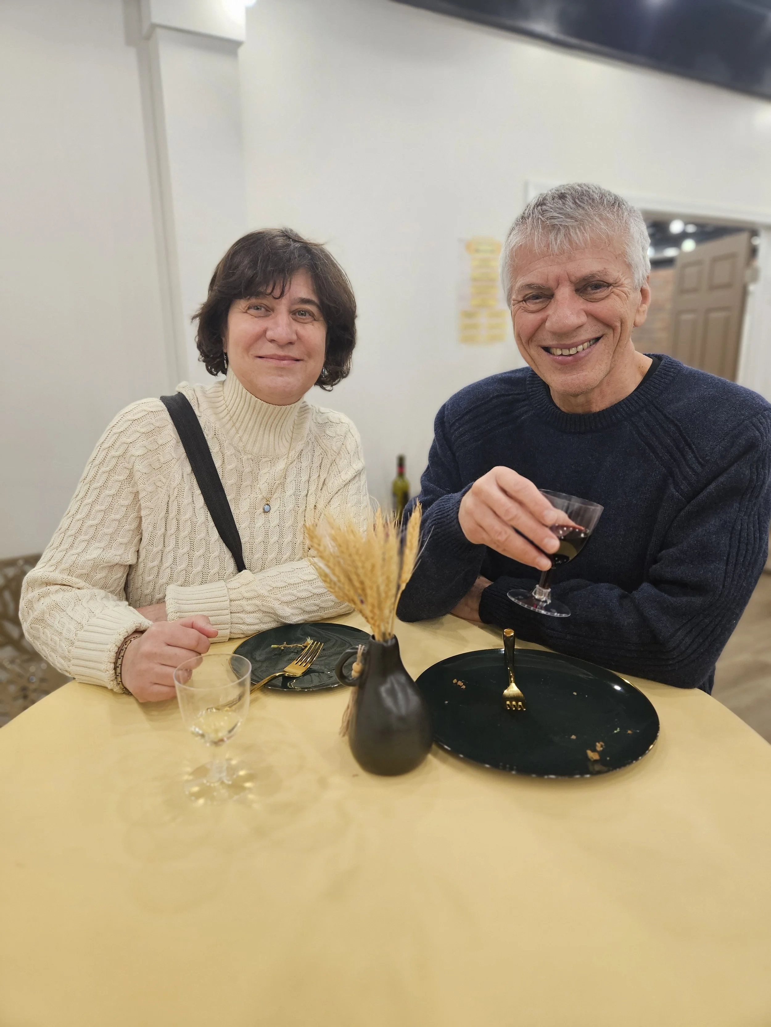 Two people sitting at a yellow table, smiling. The woman on the left has dark hair and wears a cream sweater, and the man on the right has gray hair and wears a dark sweater. There is a black plate, a glass of water, and a glass of red wine on the table. A black vase with dried wheat is in the center of the table.