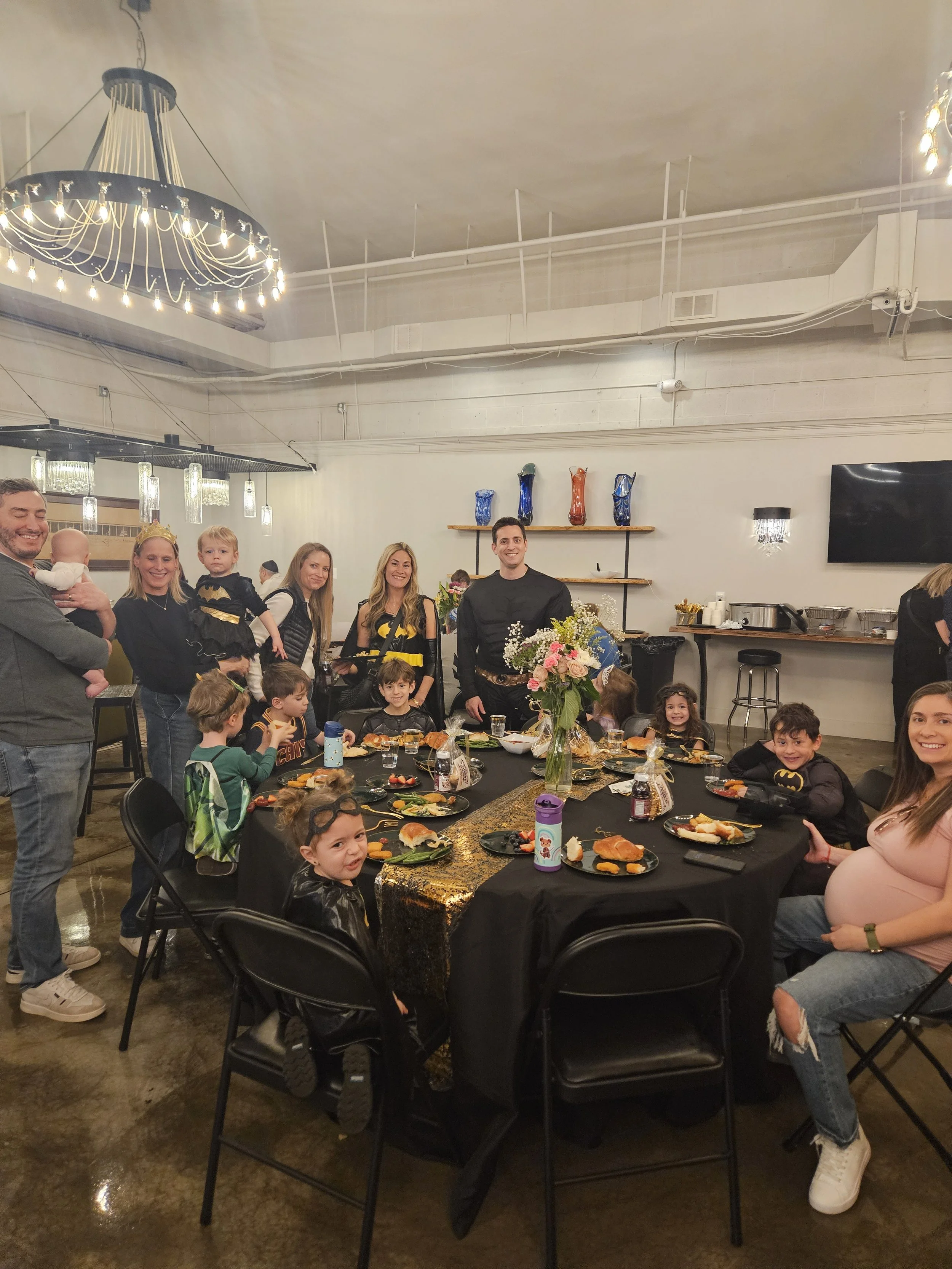 Family gathered around a decorated table for a celebration dinner, with children and adults smiling and wearing themed costumes, indoors with modern lighting and decorative vases on shelves in the background.