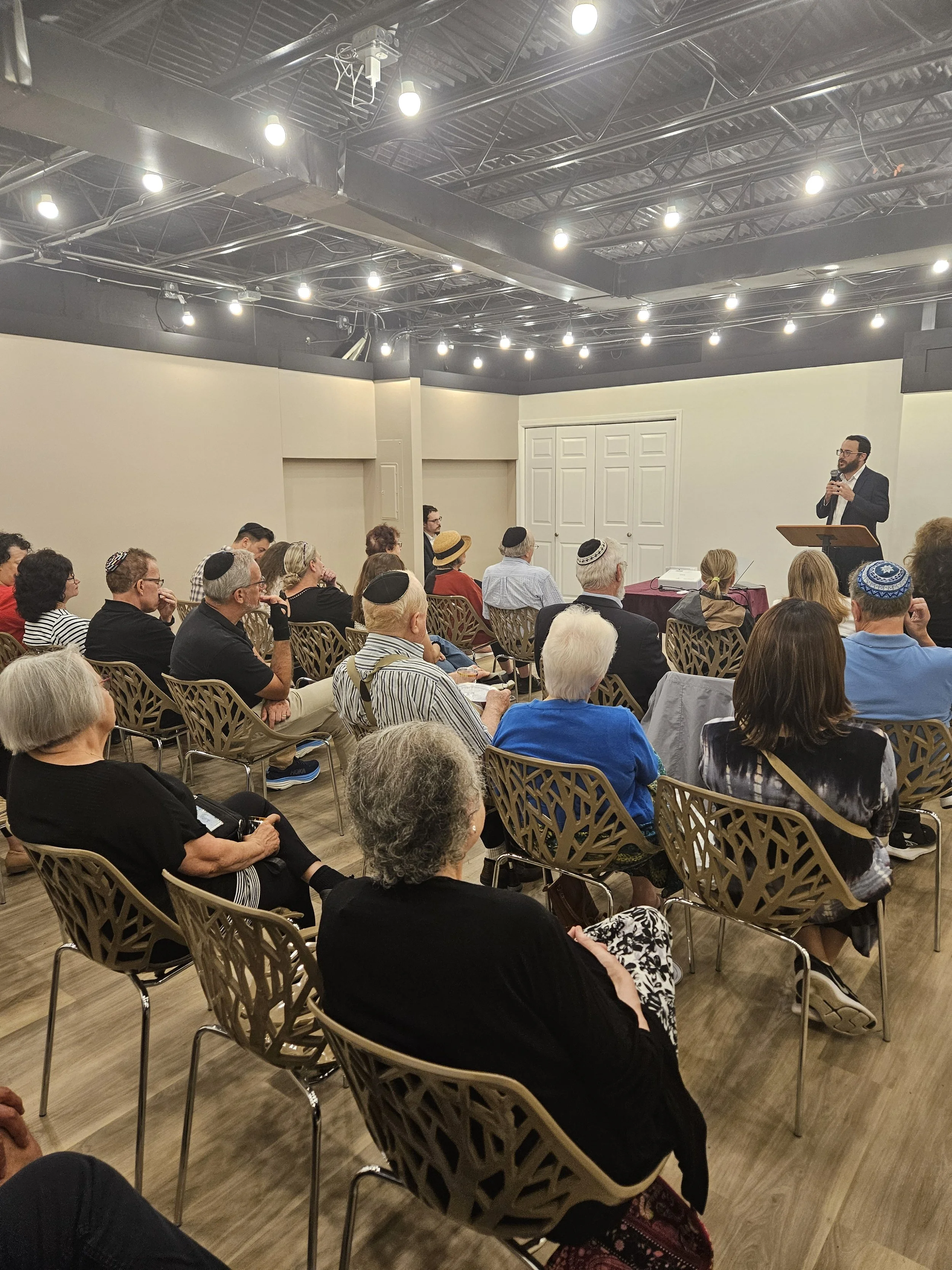 A man in a suit and glasses is speaking at a podium to an audience seated in a room with exposed ceiling pipes and string lights, in what appears to be a lecture or presentation event.
