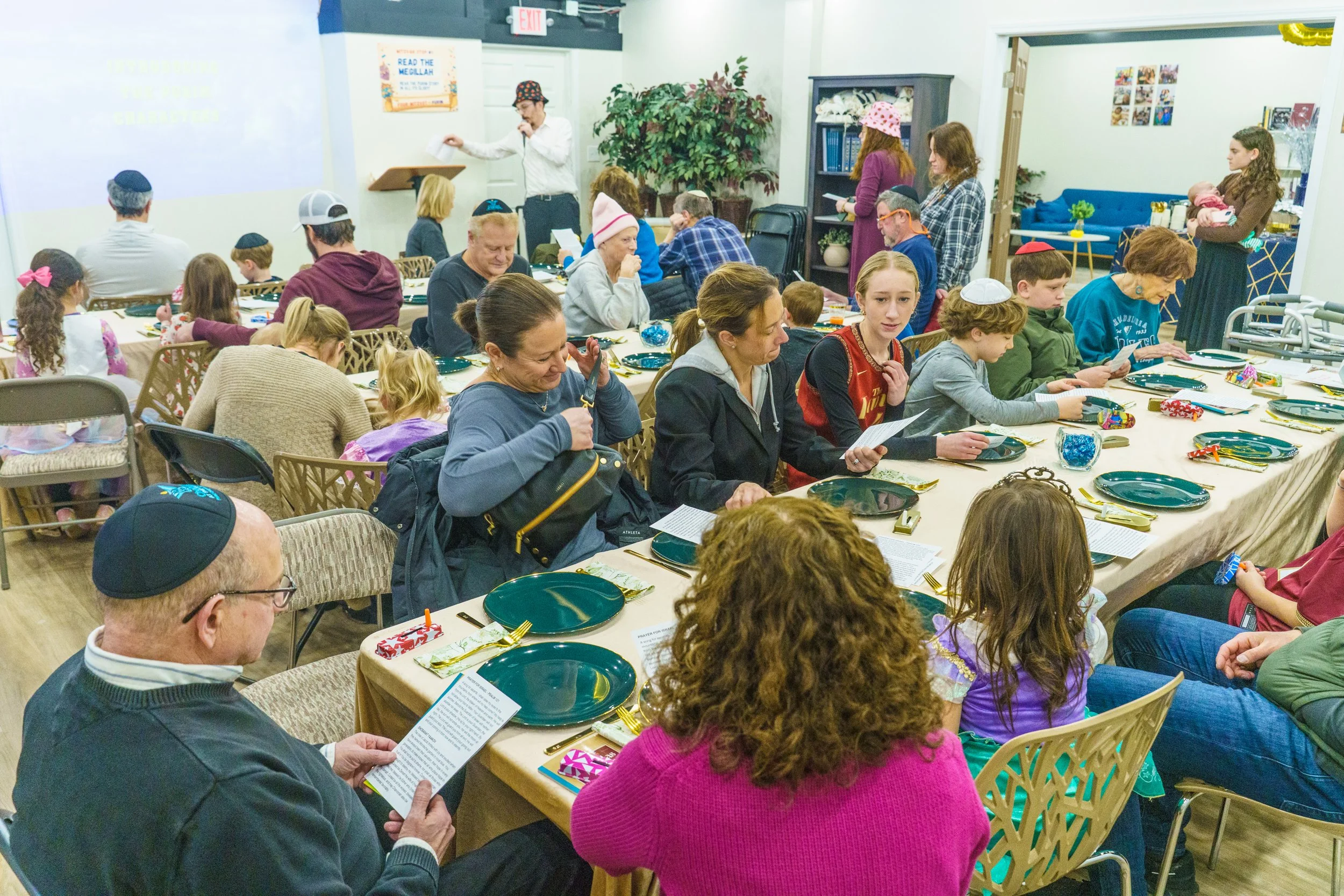 A group of people, including children and adults, gathered around a long table with plates, utensils, and party favors in a decorated indoor space. Some are reading programs, and a person is speaking at a lectern. The room has plants, bookshelves, and a projection screen.