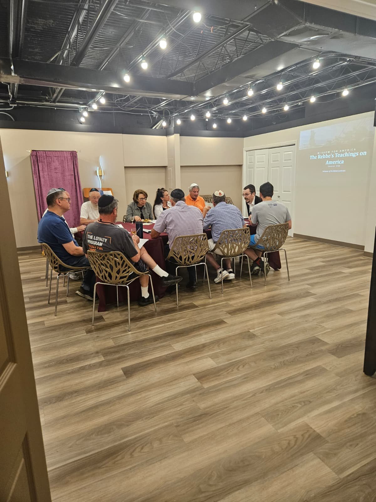 A group of people seated around a table in a conference room, engaged in discussion. The room has wood flooring and a projector screen displaying a presentation titled 'The Rebbe's Teachings on America.'