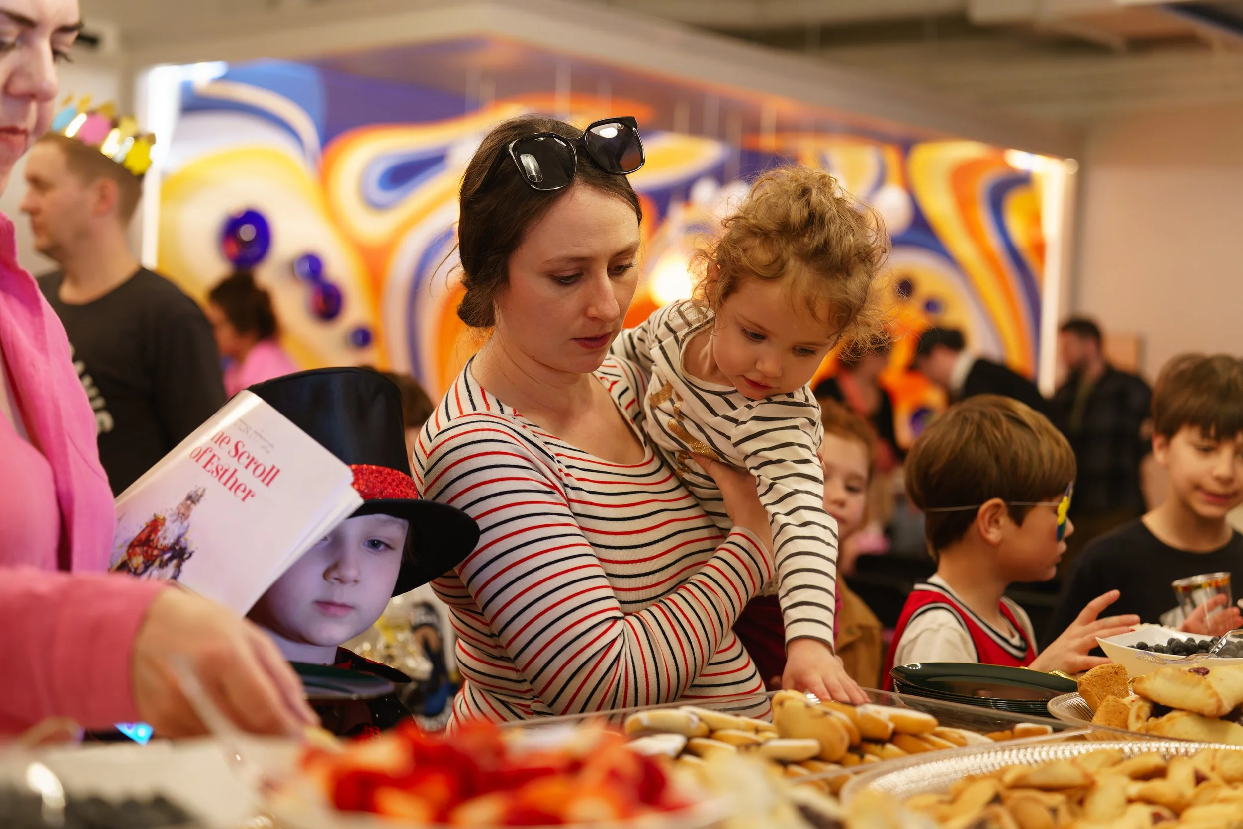 People at a table with various food items, including cookies and berries, during a festive event; a woman holding a child, with colorful abstract artwork in the background.