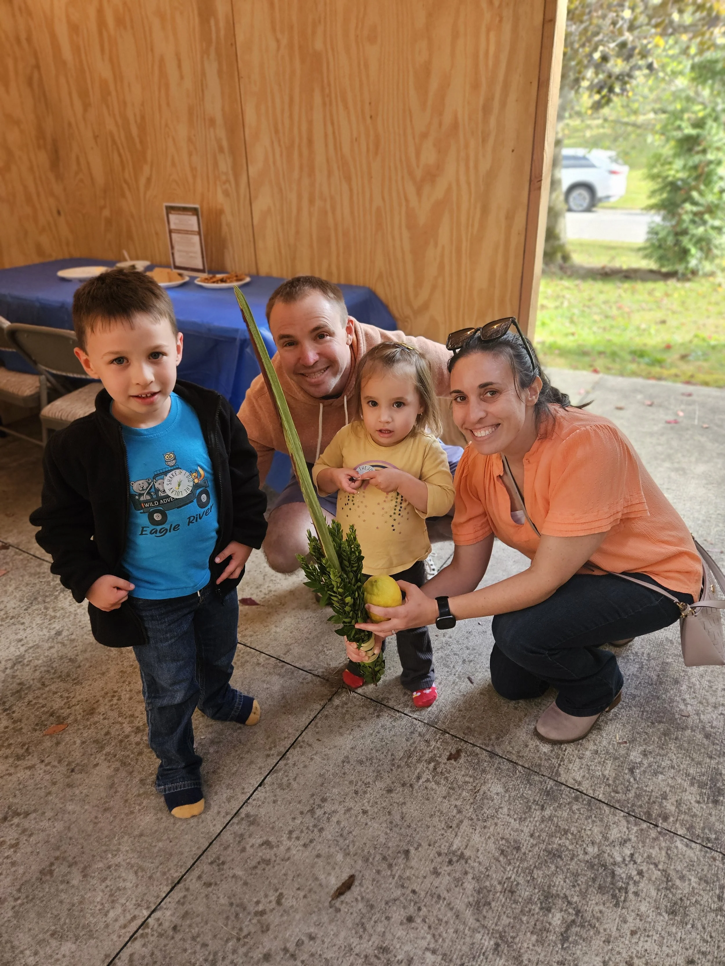 A group of four people, including two children and two adults, smiling and crouching outdoors under a wooden structure. A woman is holding a plant and an apple, with all four smiling at the camera. There's a table with snacks in the background near a wood-paneled wall.