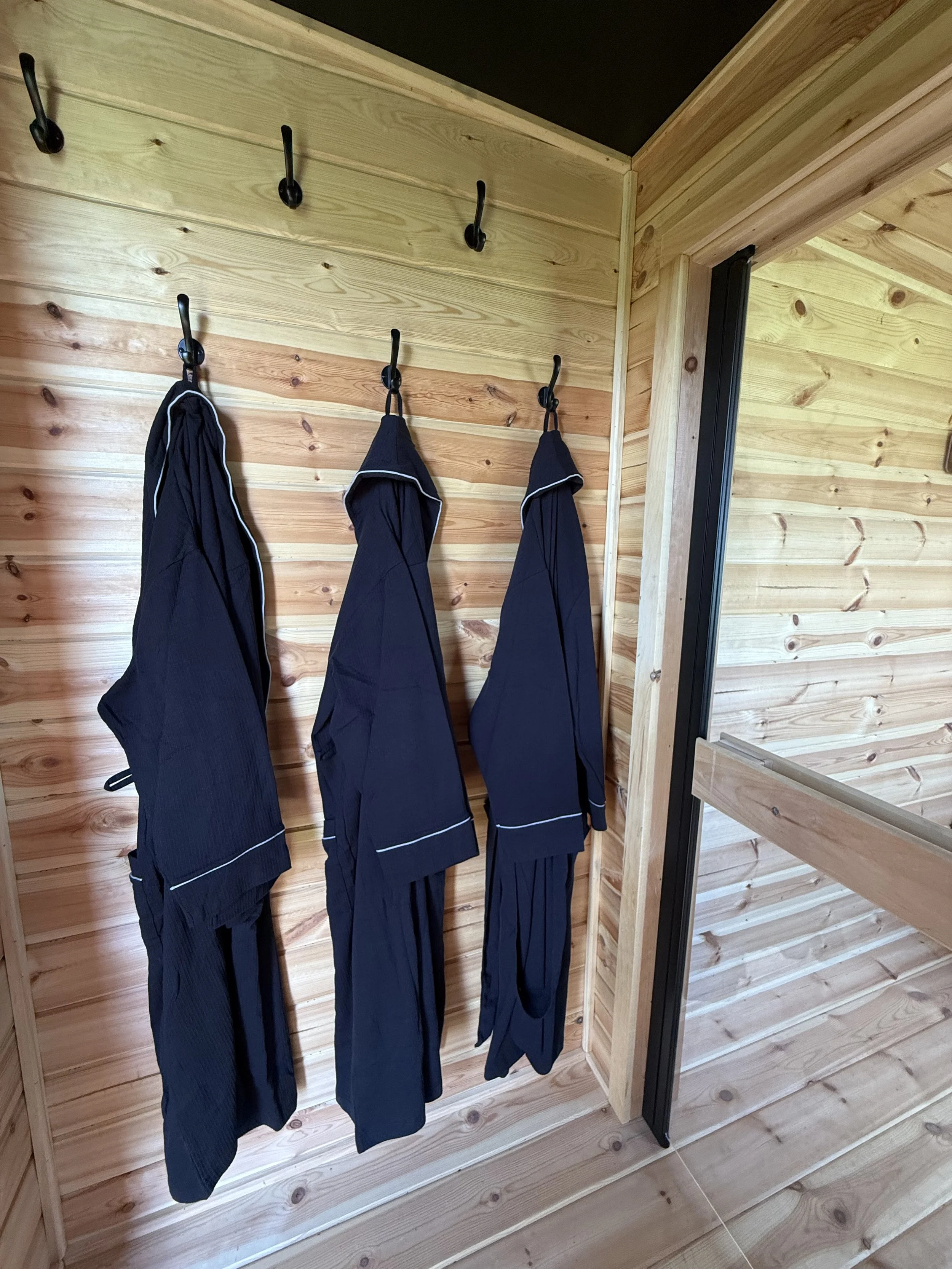 Three black bathrobes hanging on black hooks on a wooden wall inside a room with wooden flooring and walls.