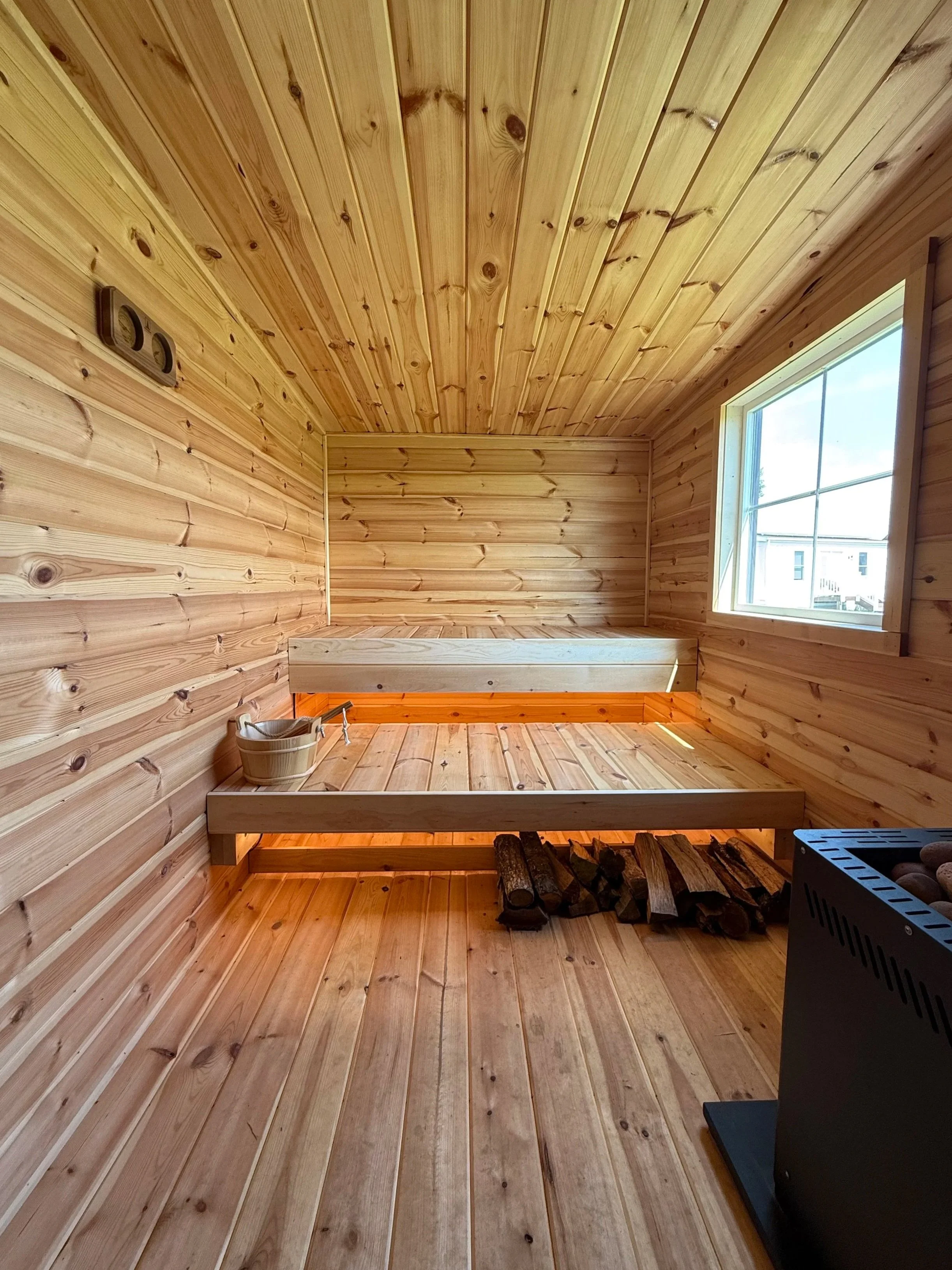 An interior view of a small sauna room with wooden paneling on the walls, ceiling, and floor, a window on the right, a wooden bench along the back wall, a bucket with a ladle on the left, neatly stacked firewood on the floor, and a heater at the bottom right corner.
