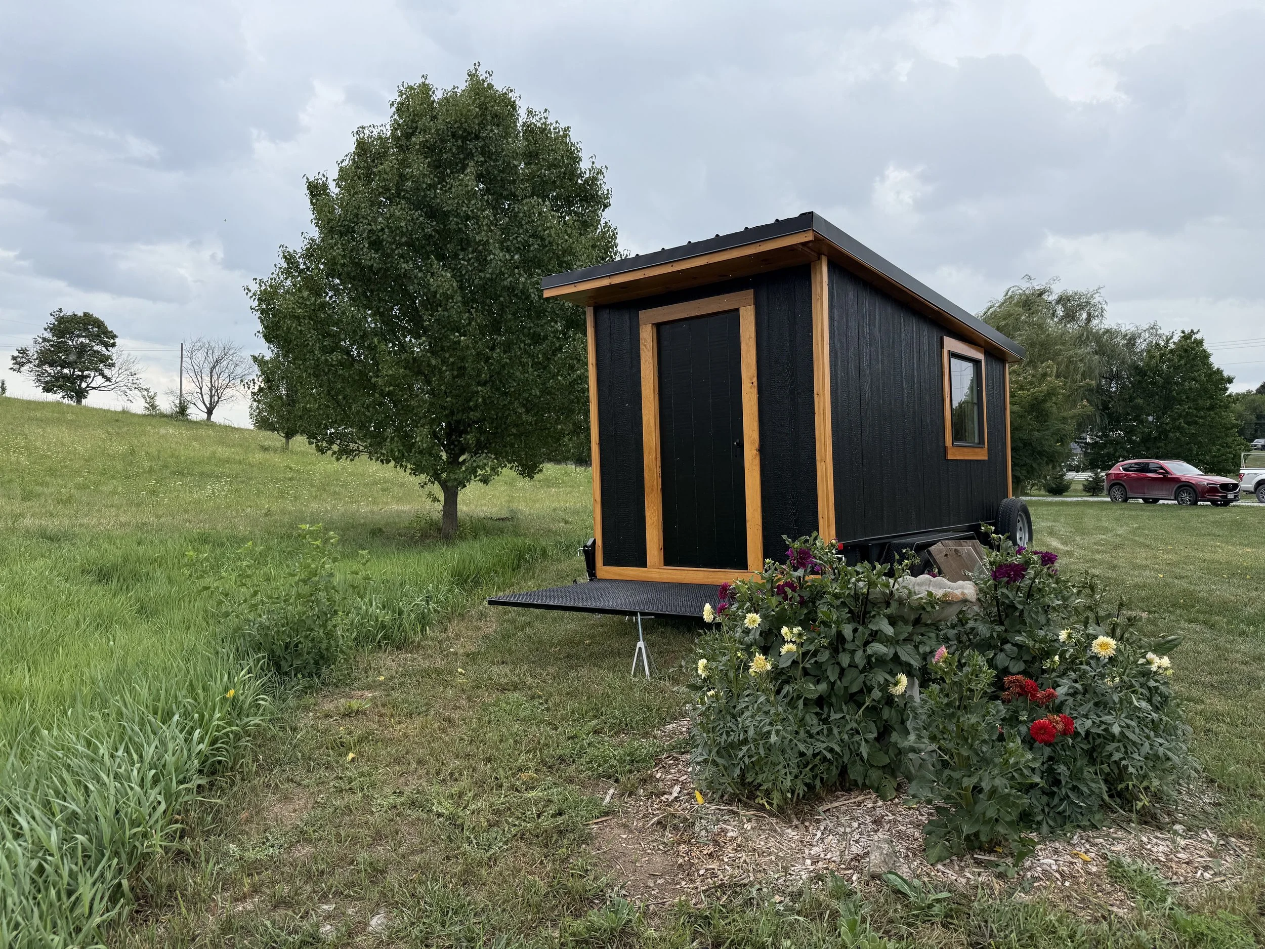 A black mobile sauna on wheels in a grassy field, with trees and bushes in the background under a cloudy sky.