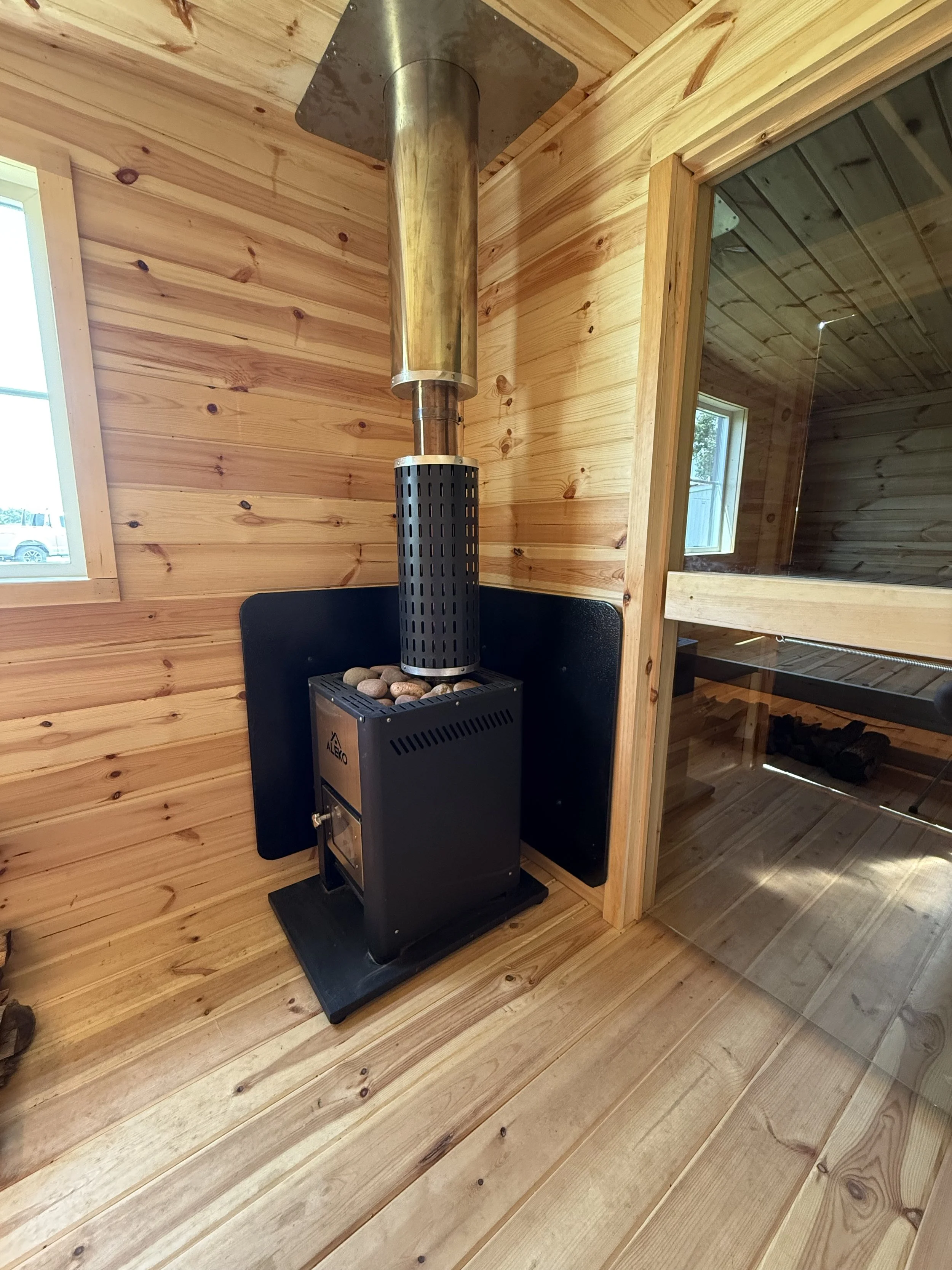 A wood-burning stove with a metal chimney pipe in a wooden sauna room, with wood-paneled walls and floors, and a glass door leading to a sauna or steam room.