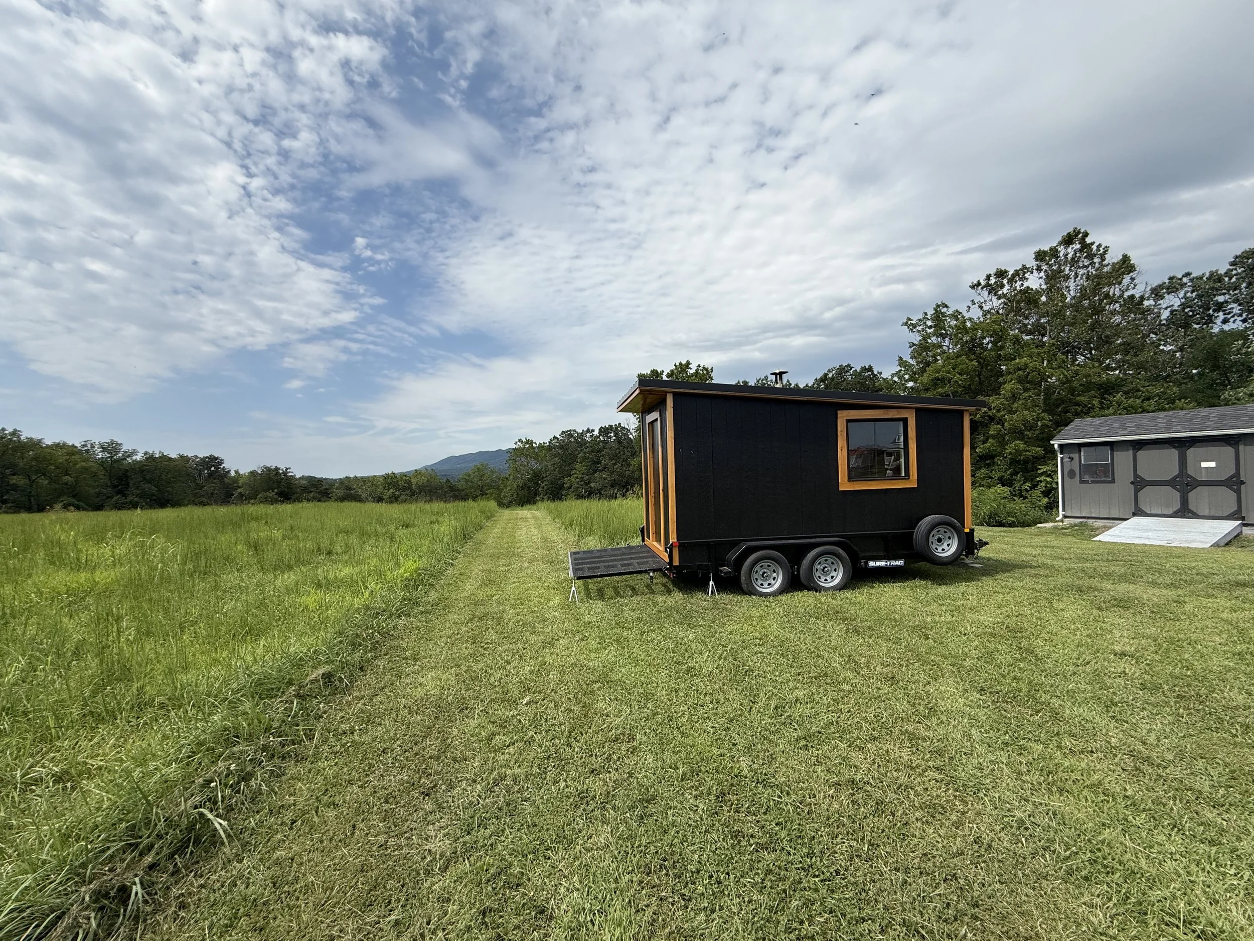 A small black mobile sauna on a trailer with a wooden window frame, set on a grassy field with a gravel path, surrounded by trees and hills under a partly cloudy sky.