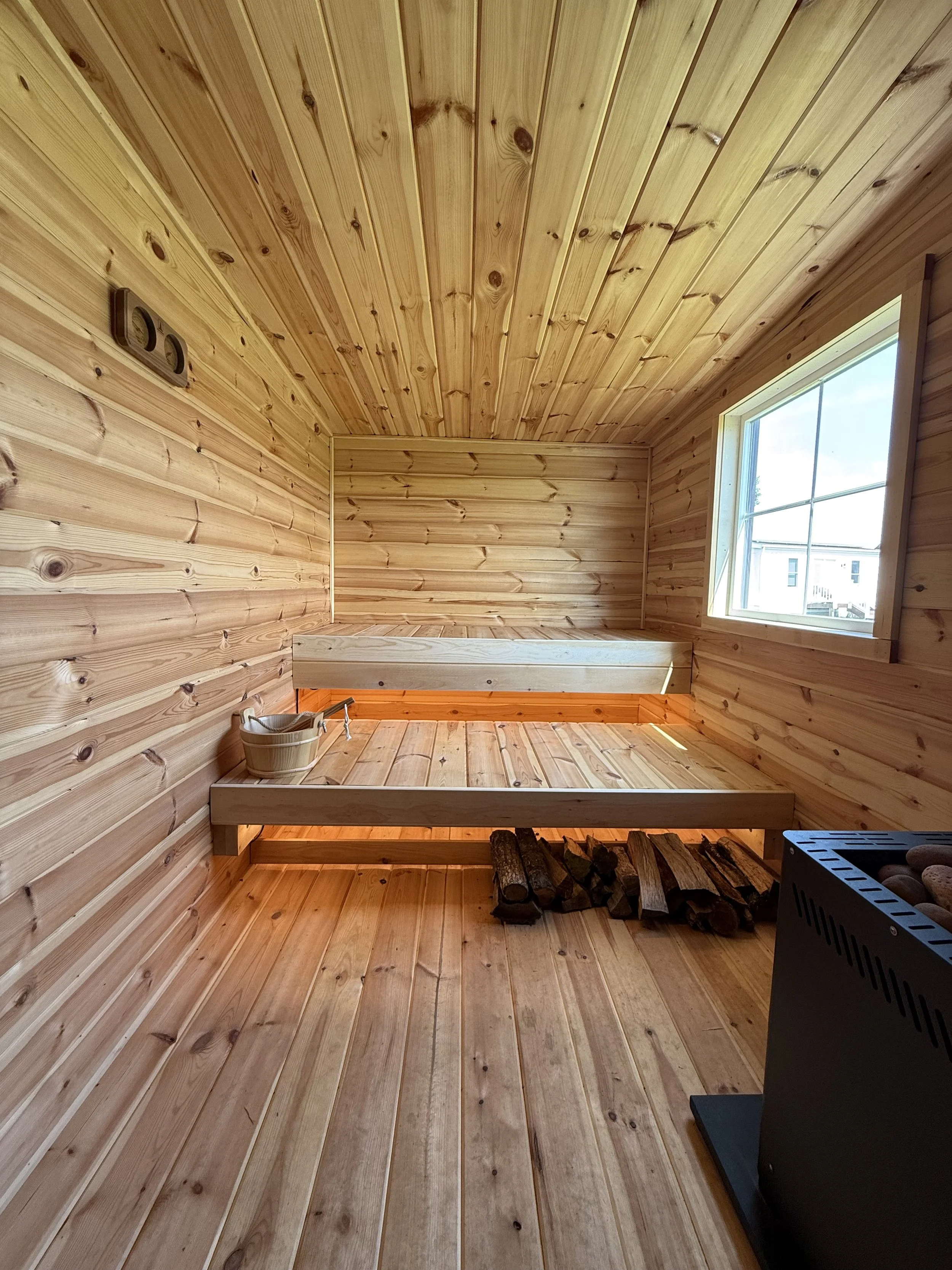 Inside a cozy wooden sauna with wood-paneled walls, ceiling, and floor. There is a window on the right side, a built-in wooden bench, a bucket with a ladle, a stack of firewood, and a heater with stones.
