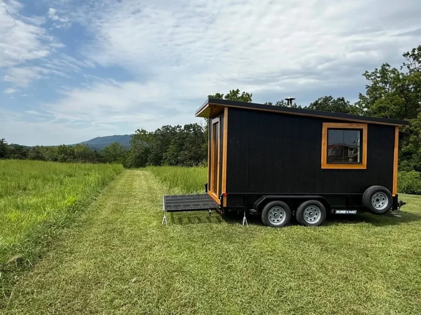 A mobile sauna on a trailer with a front porch and window, set in a grassy field with trees and mountains in the background, under a cloudy sky.
