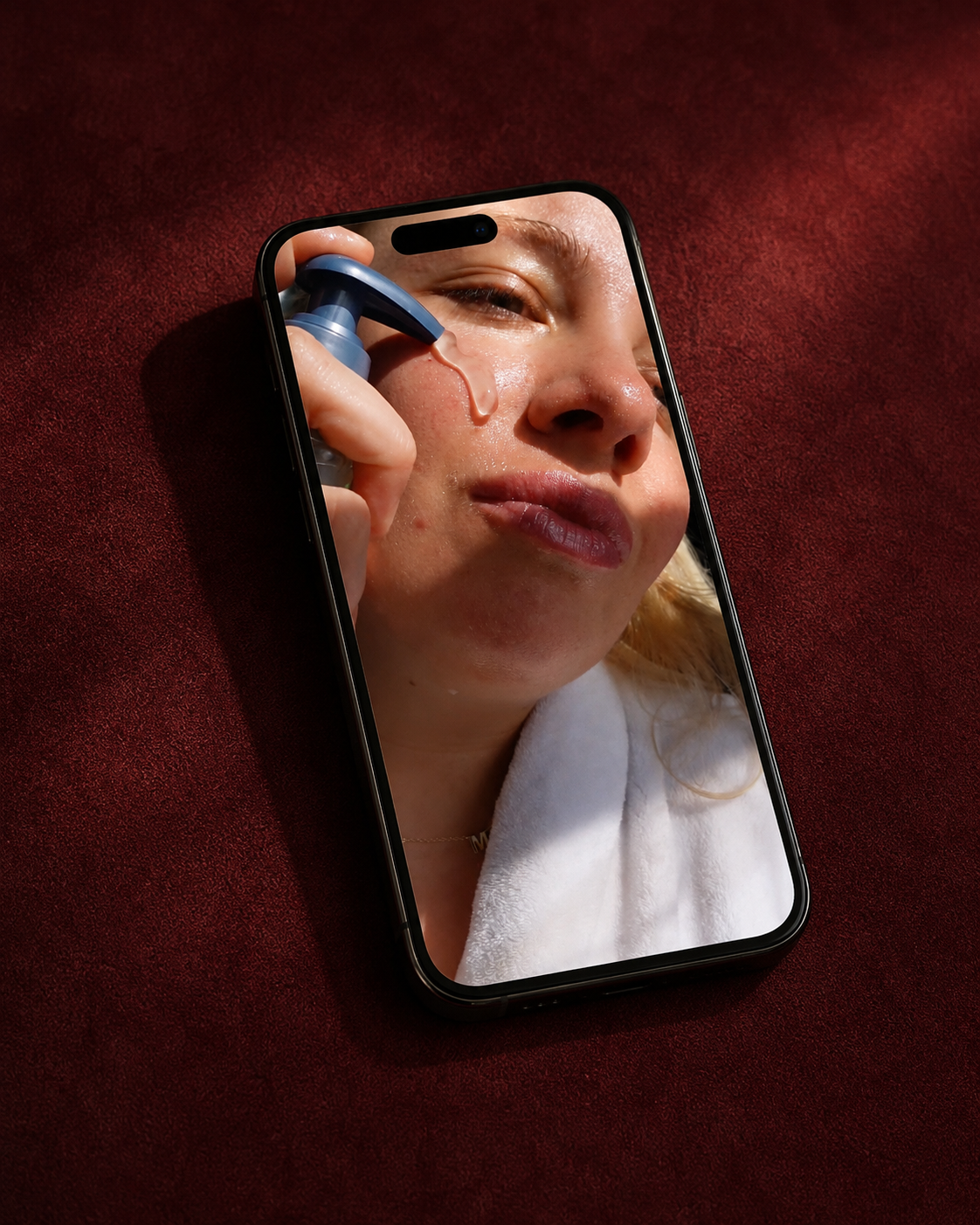 A close-up of a woman looking at her phone, which displays an image of her applying skincare product around her nose.