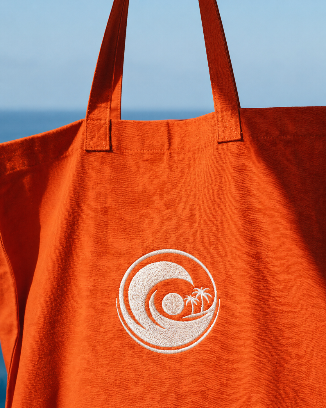 Orange tote bag with white circle logo featuring a sun, waves, and palm trees, outdoors with a blue sky and ocean in the background.
