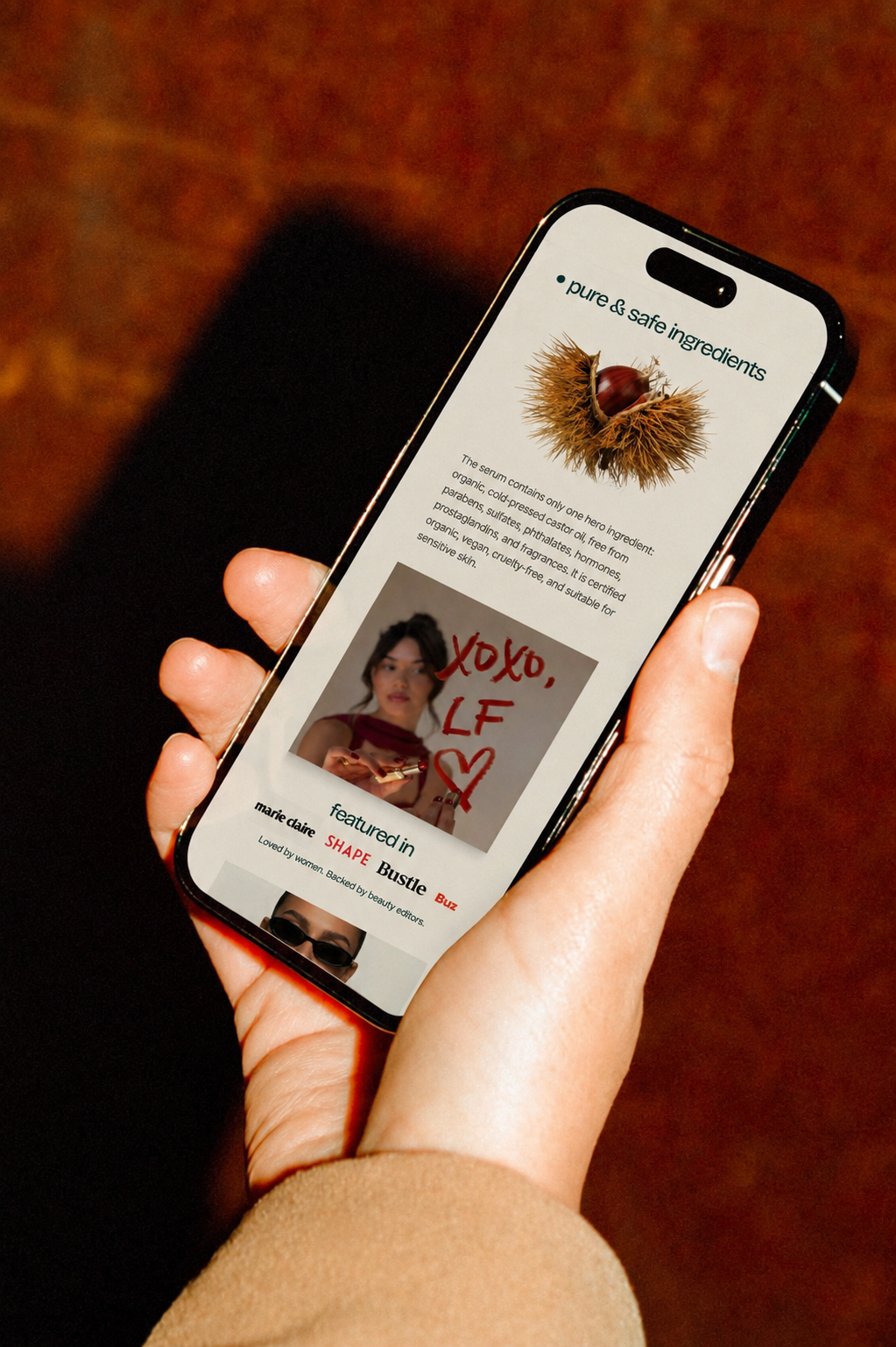 A person holding a smartphone displaying a webpage about skincare ingredients and features. The webpage highlights pure and safe ingredients, showing a chestnut image, a photo of a woman with handwritten "XOXO, LF" message, and mentions several magaz