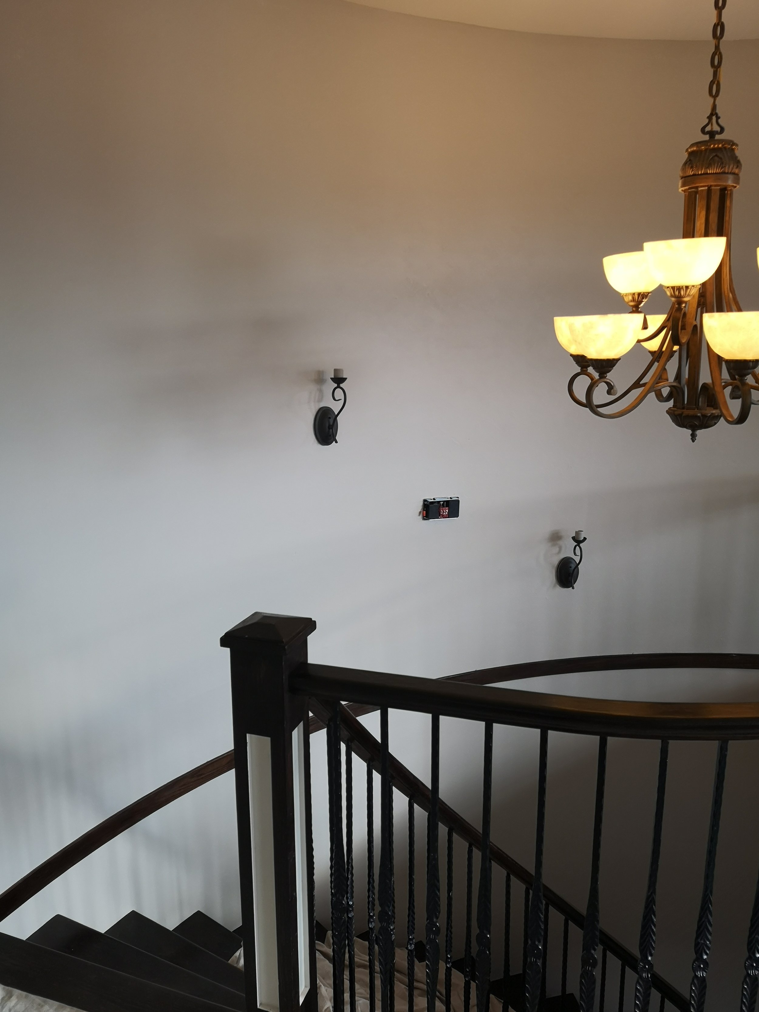 Interior view of a staircase with black railings and dark wood handrail, a chandelier with multiple glowing shades, a beige wall, wall-mounted lamps, and a device on the wall.