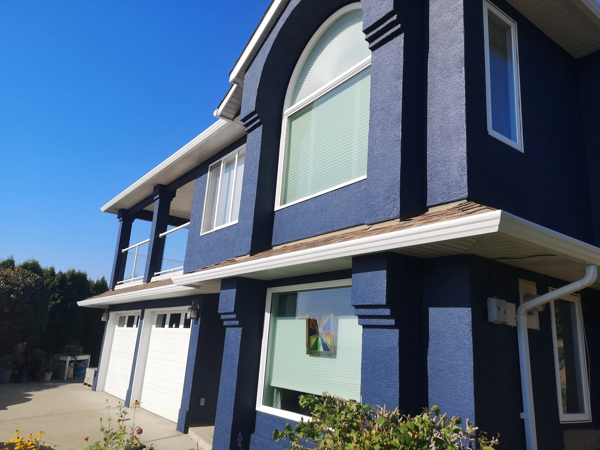 A blue house with white trim, large arched window, smaller rectangular window, and a white garage door, with a driveway and garden plants in front, under a clear blue sky.
