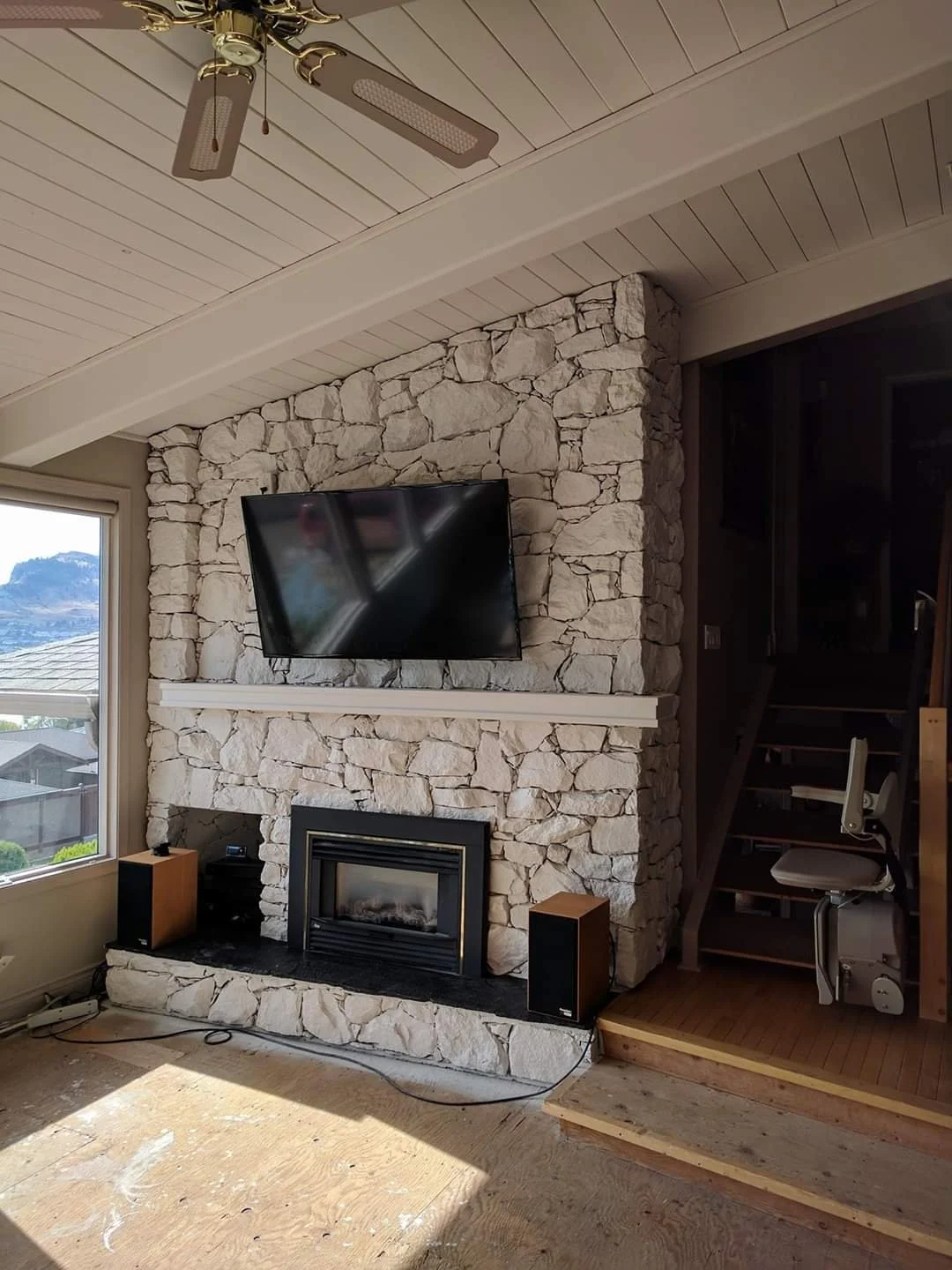 Living room with a stone fireplace, a mounted flat-screen TV, a window showing a view of houses and a mountain, and a ceiling fan with lights.