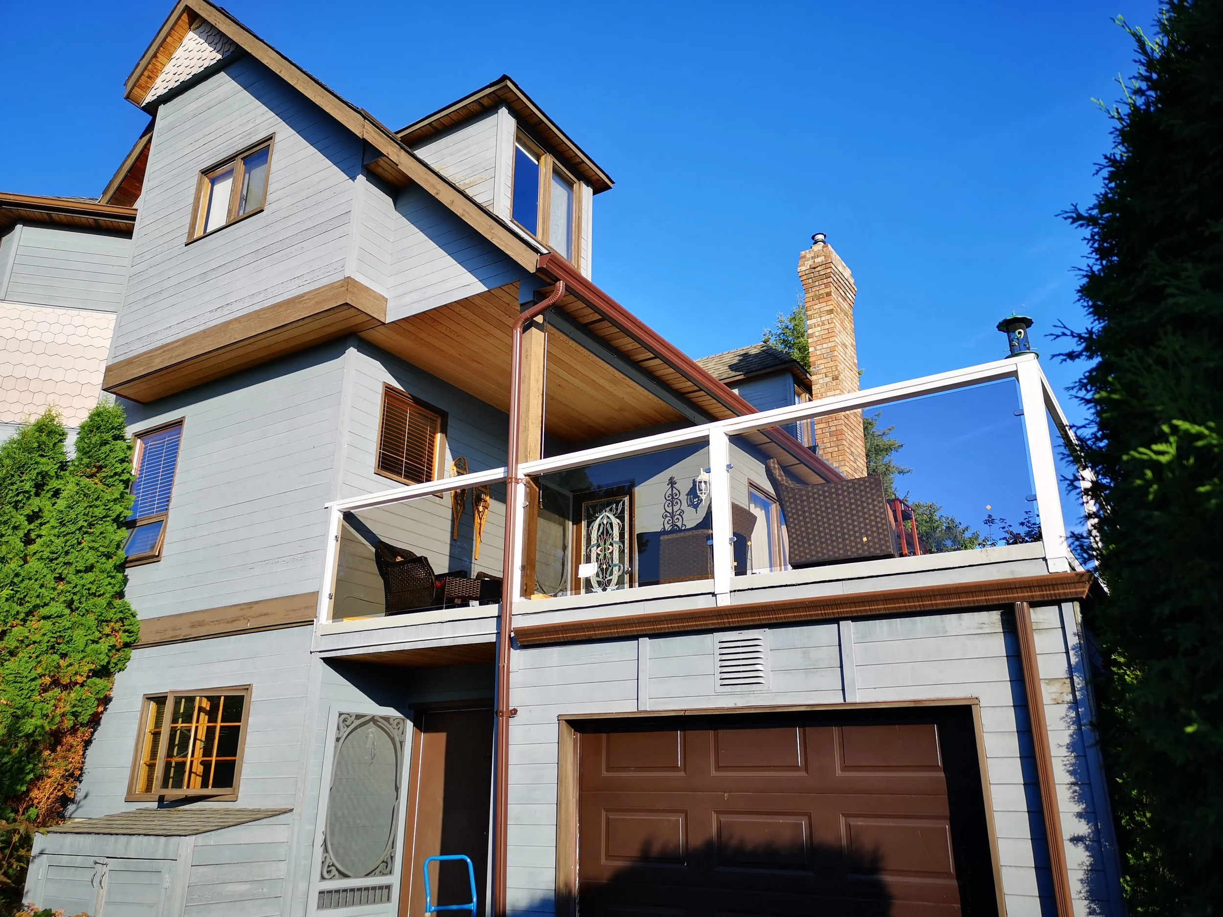 A multi-story house painted in light blue with brown accents on the windows and garage door. It features a balcony with a white railing, a brick chimney, and large windows. The house is under a clear blue sky, with green trees on the right and left sides.