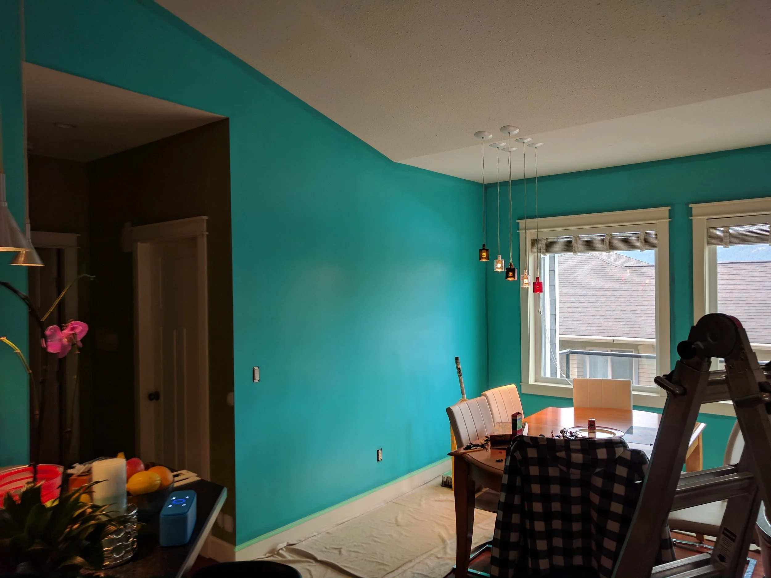 A dining area with a teal accent wall, a white ceiling with modern pendant lights, a window with blinds, and a table with chairs. Some items and a window view of rooftops are visible.