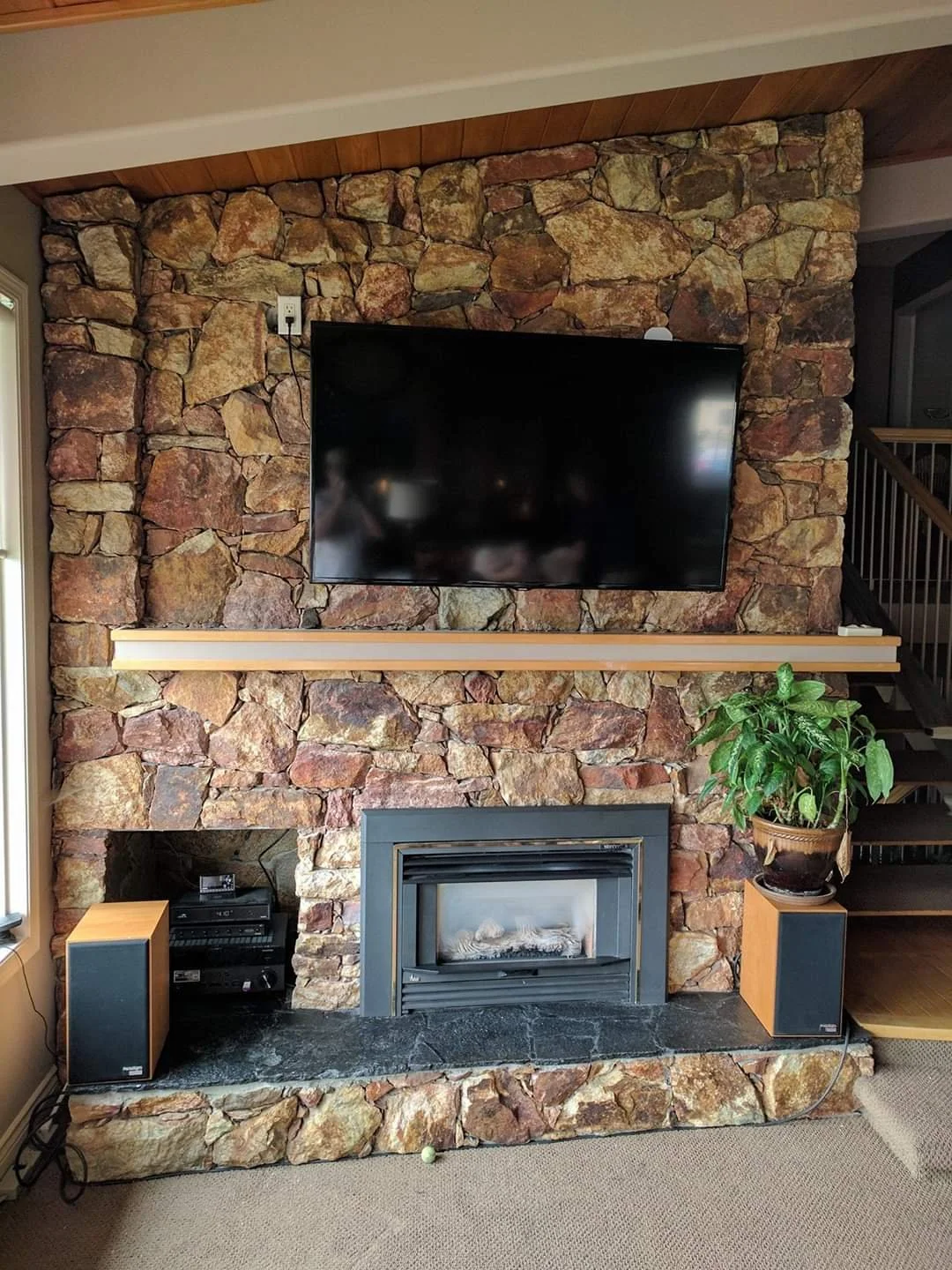 Living room with a stone fireplace, a mounted flat-screen TV, a potted plant on a side speaker, and audio equipment on the hearth.