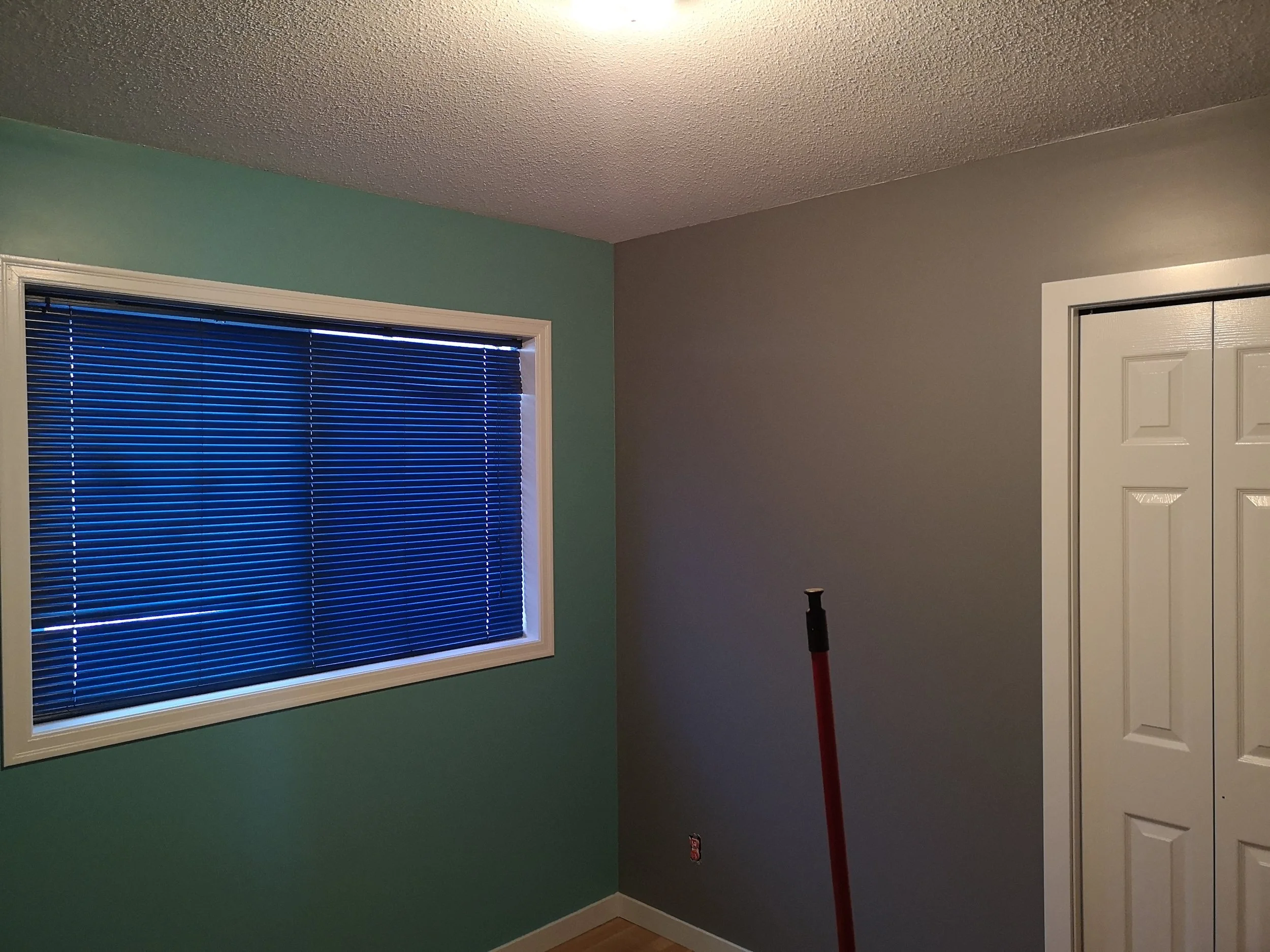 A room corner with light green and beige walls, a window with closed blue blinds, and a white door. A red broom leans against the wall.