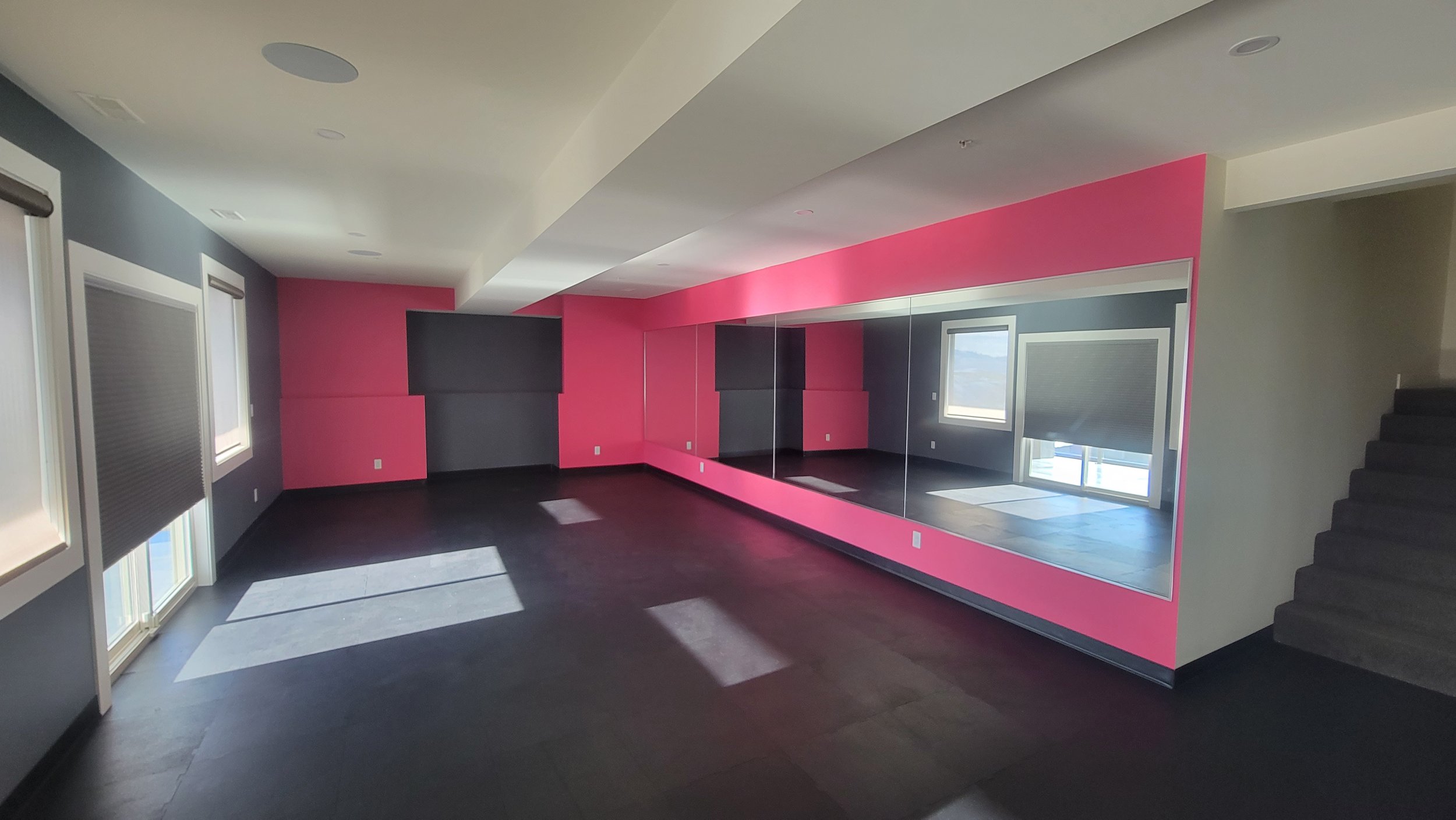 Empty dance or exercise studio with dark flooring, pink and gray walls, large mirrors, windows with blinds, and a staircase to the right.