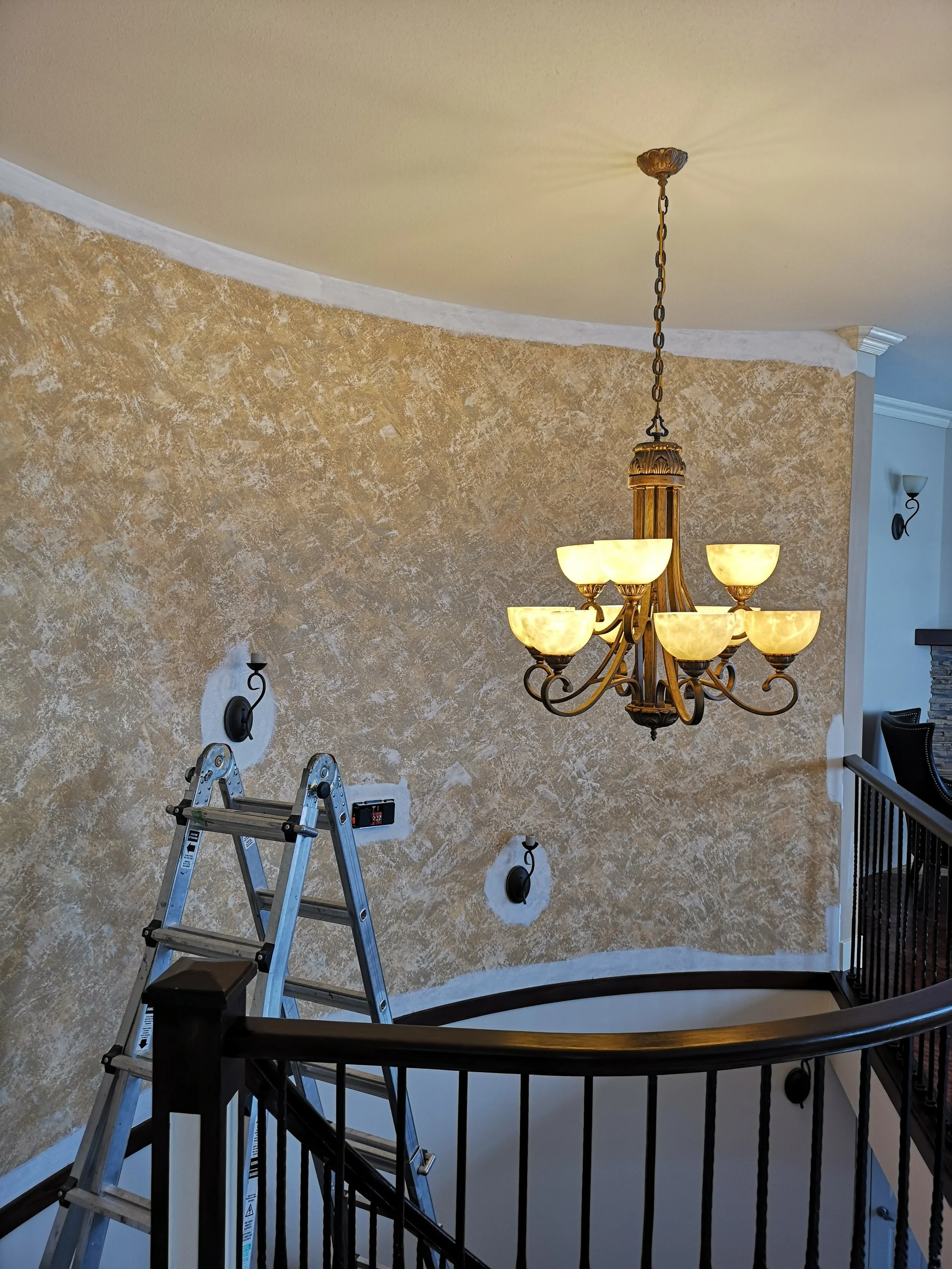 Interior view of a staircase with a chandelier hanging from the ceiling, a ladder in the foreground, and a textured beige wall.