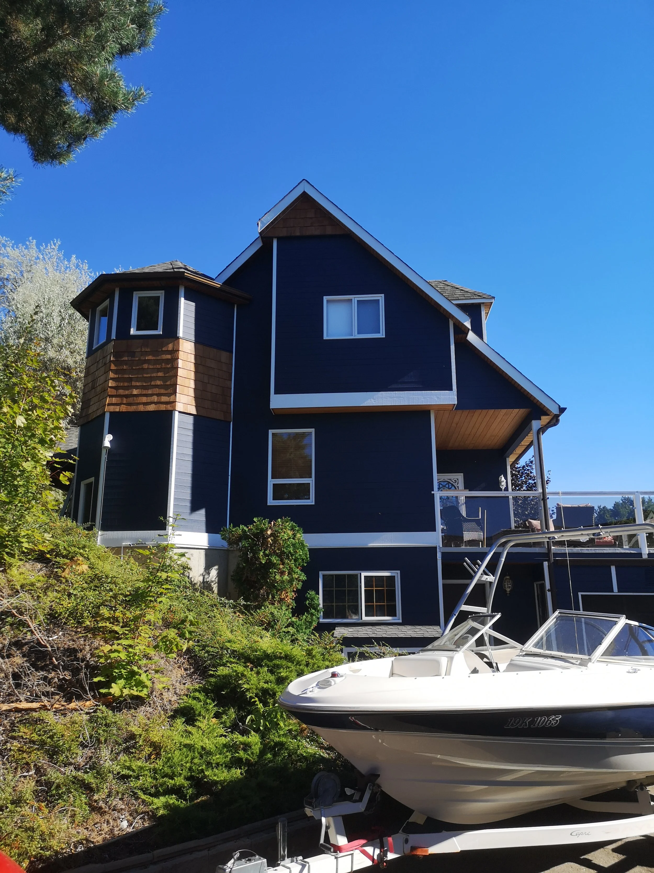 A multi-story house painted dark blue with white trim, situated on a hill. The house features window bays, a sloped roof, and a balcony. In front of the house, there is a white boat on a trailer, parked on the driveway. The background includes a clear blue sky and greenery with trees and bushes.